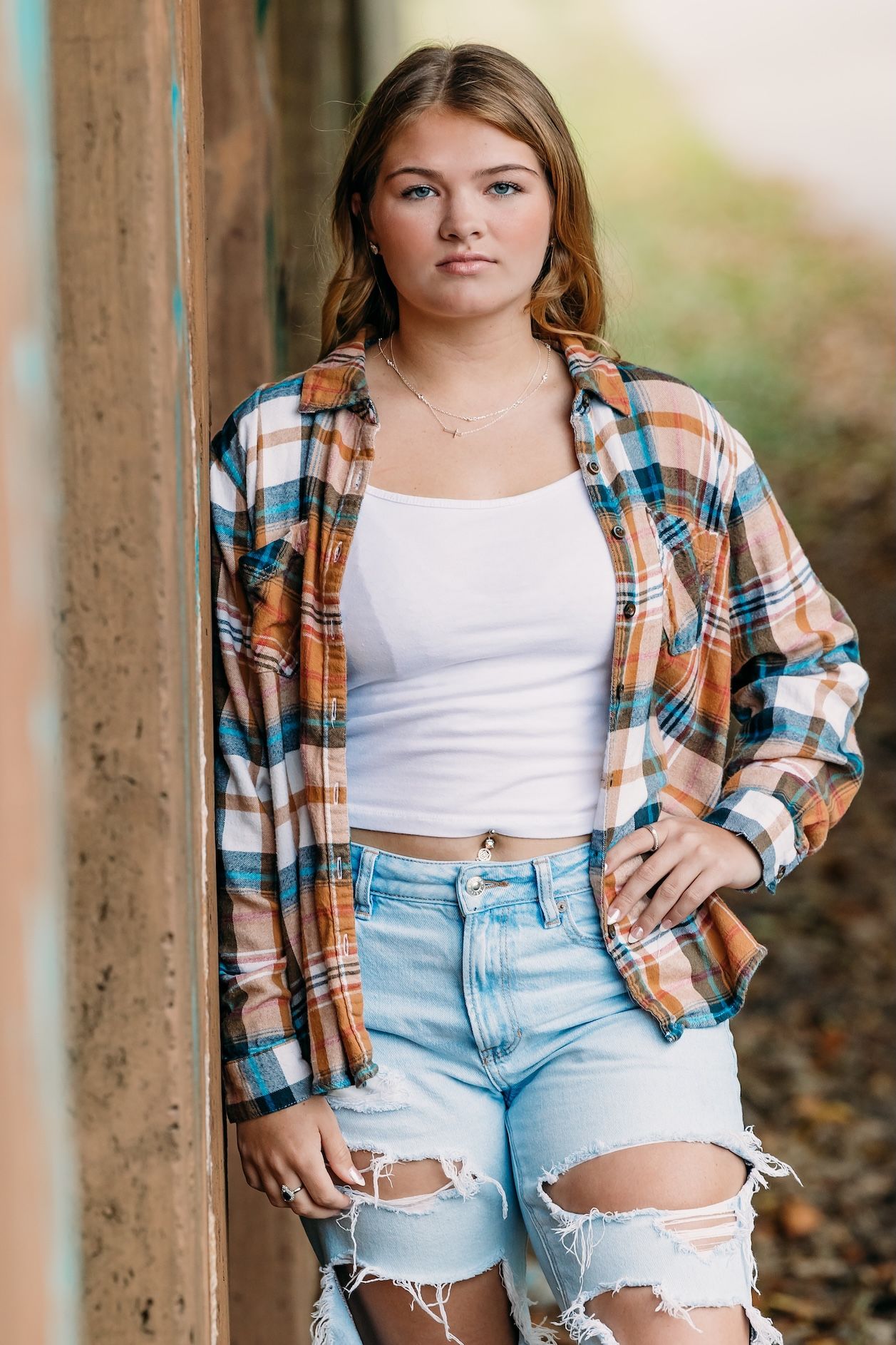 A person wearing a plaid flannel shirt, white tank top and distressed jean shorts poses against a wooden post outdoors.