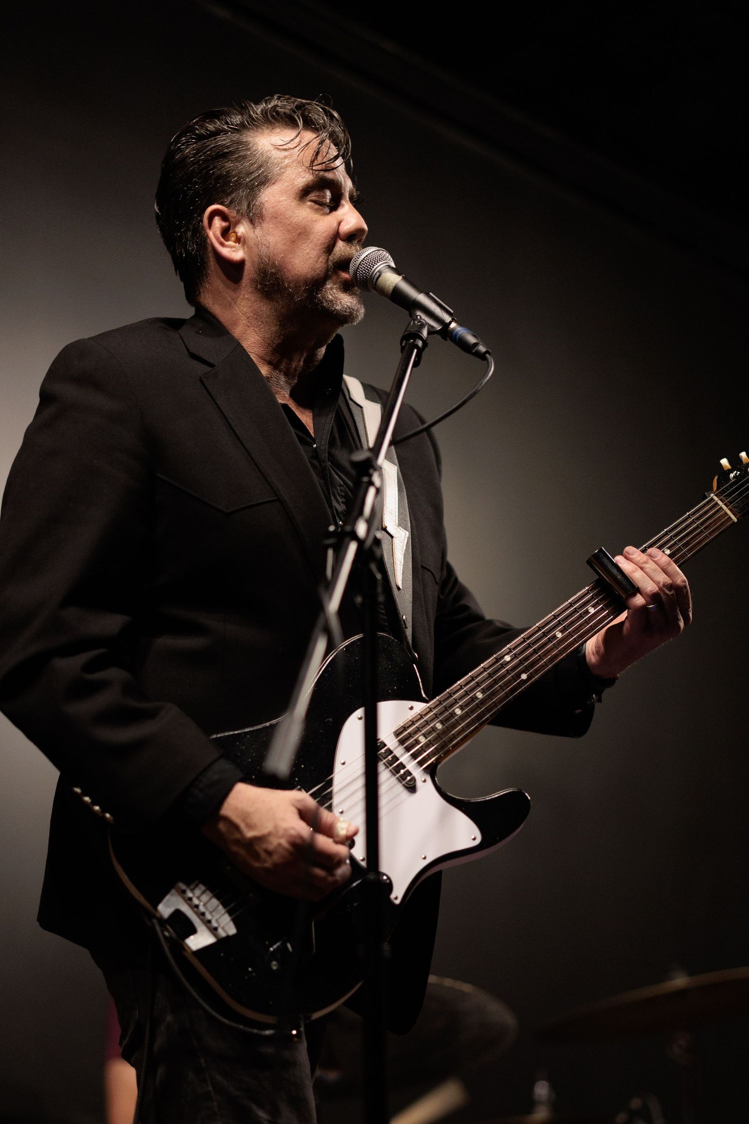 Musician in black suit jacket performs passionately on stage with bass guitar and microphone in dramatic lighting.
