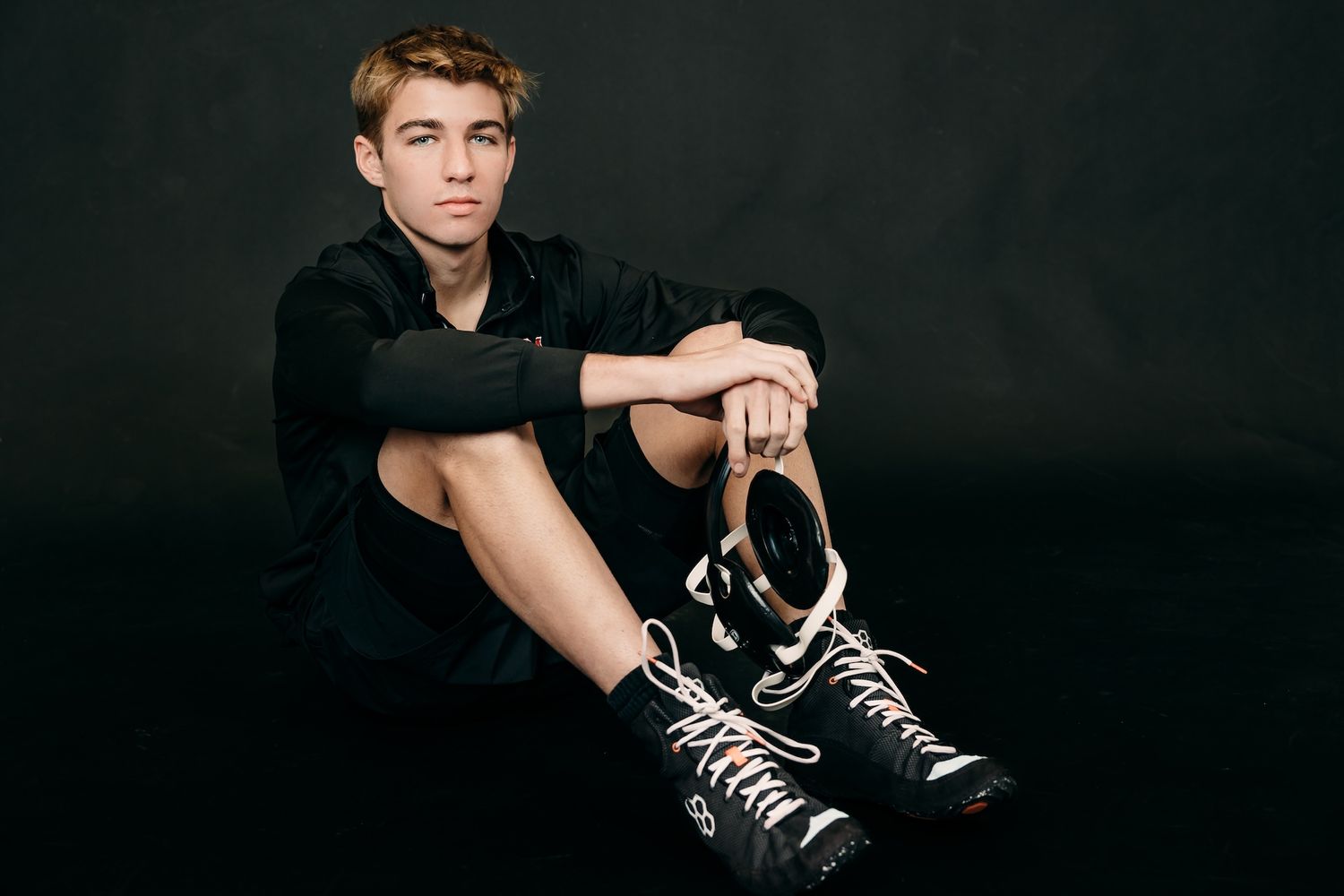 Portrait against dark background of someone wearing a black shirt and white and black sneakers sitting on floor.