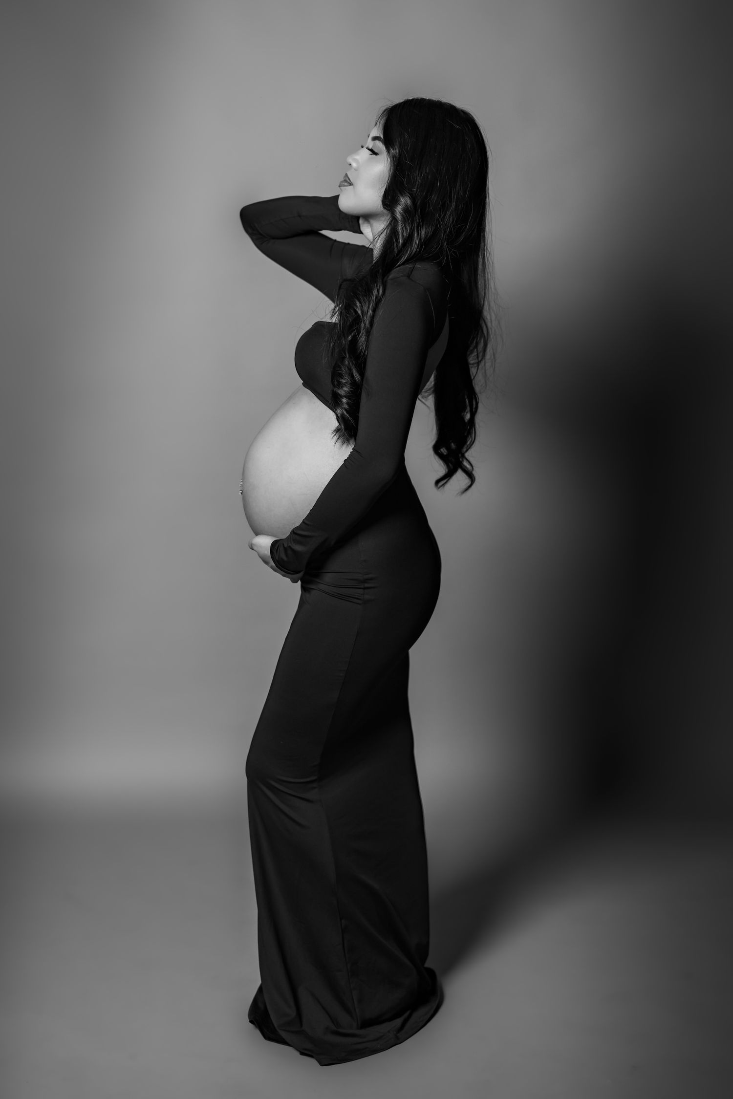 Black and white maternity portrait of a woman in a long dark dress posing elegantly in profile against a gray backdrop.