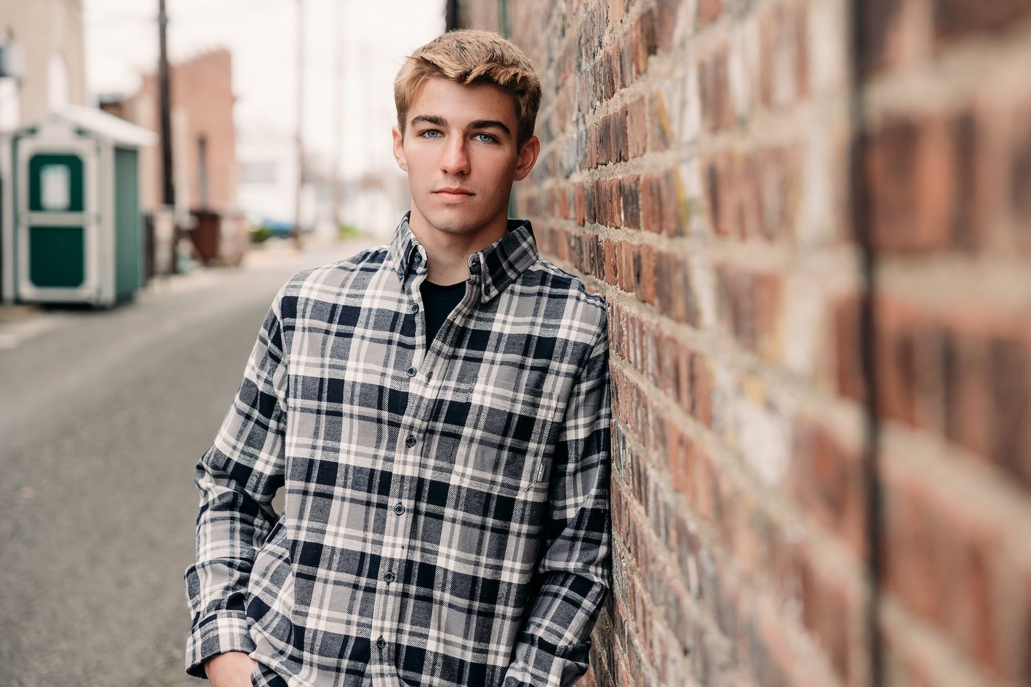 Stylishly dressed person in plaid flannel shirt leaning against brick wall on urban street.