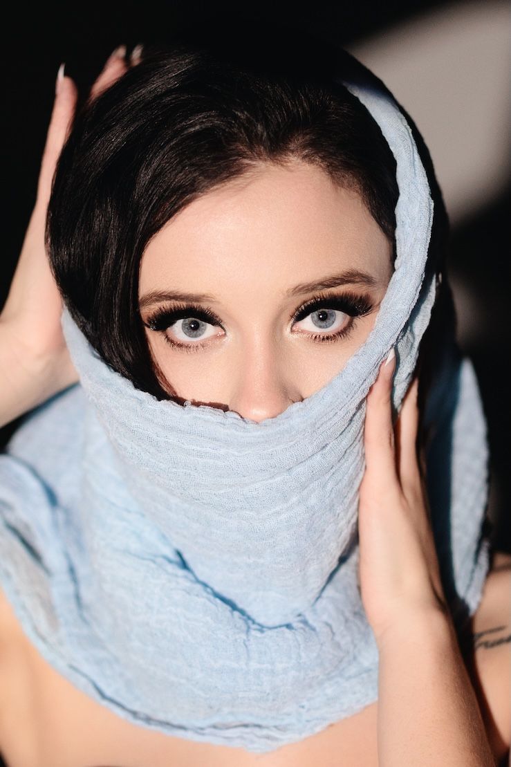 Person wearing a light blue scarf wrapped around their head with striking eye makeup looking at camera.