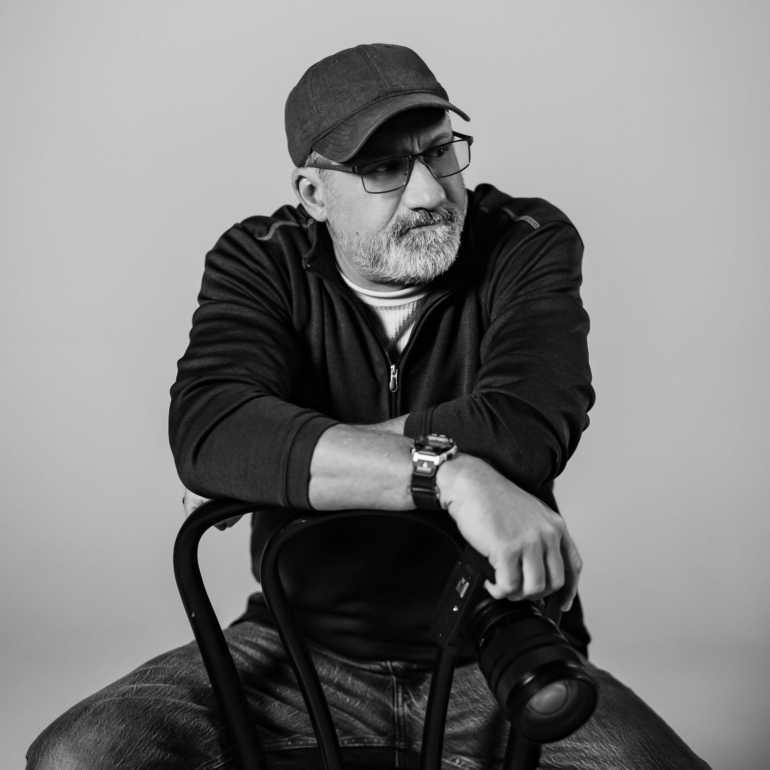 Black and white portrait of a mature person wearing glasses, baseball cap and hoodie sitting casually in a chair.