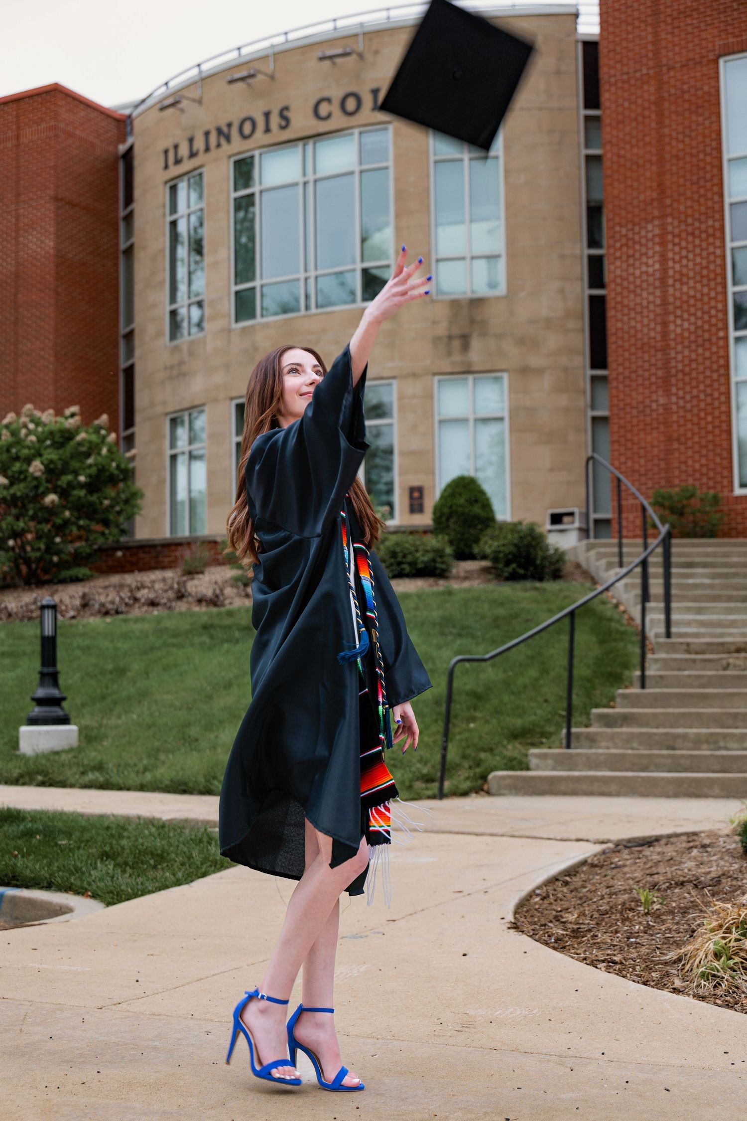 Graduate in cap and gown celebrates by throwing mortarboard in front of Illinois College building on graduation day.