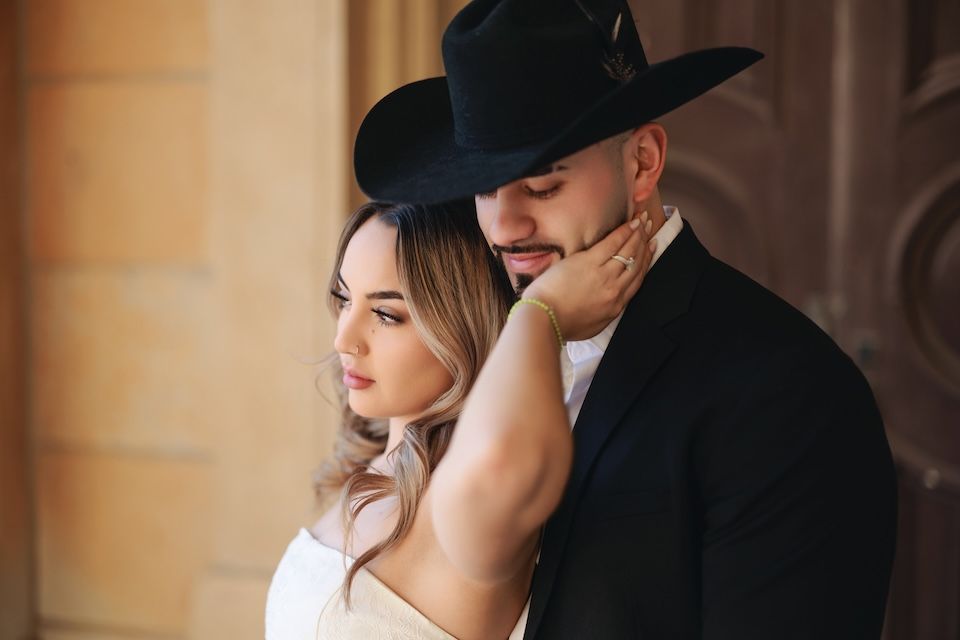 A romantic couple in western style attire share an intimate moment against a rustic wall.