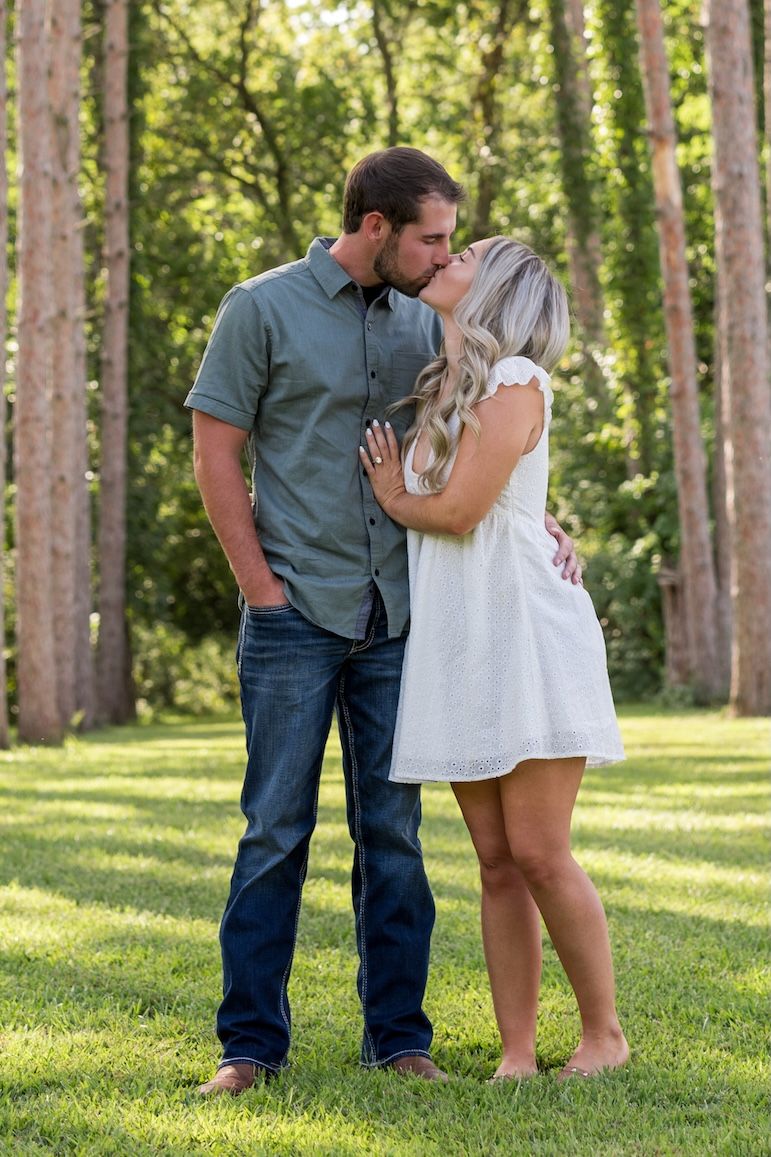 A couple shares an intimate moment while standing together on grass in a sunlit forest setting.