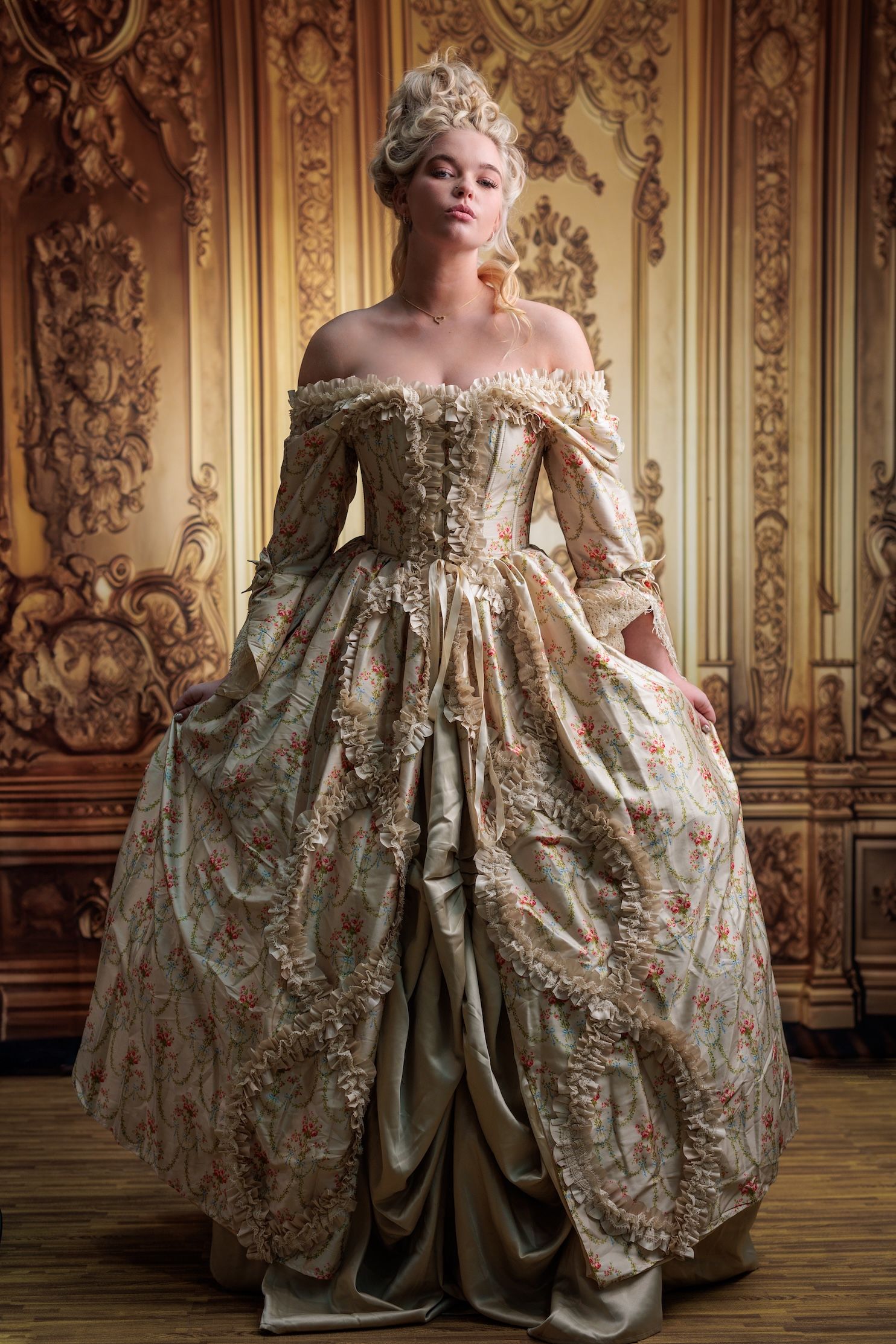 A woman wears an ornate 18th century rococo-style brocade ball gown in a gilded palace room with decorative wall panels.