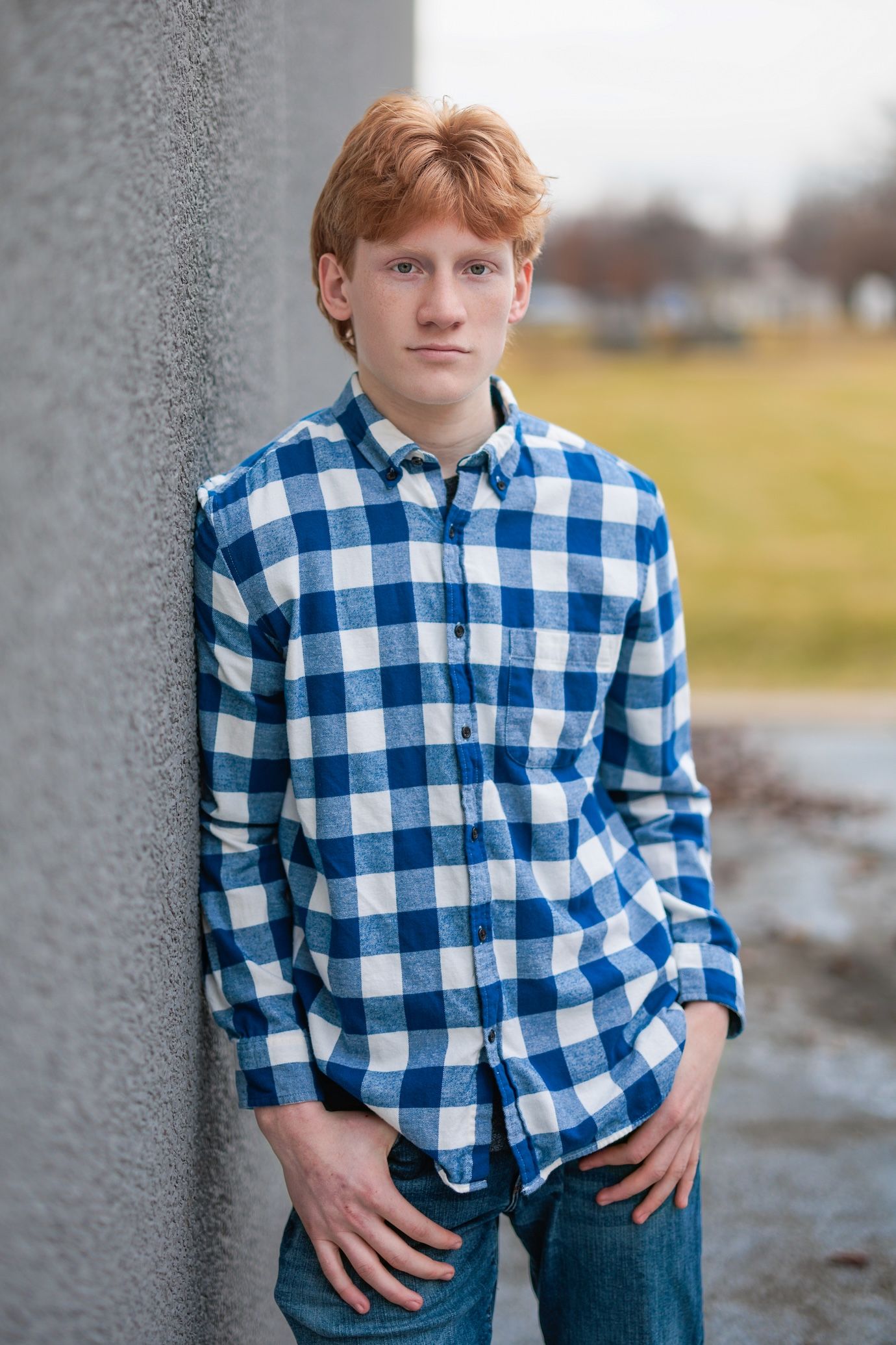 Person wearing blue and white checkered button-down shirt leaning against gray wall outdoors.