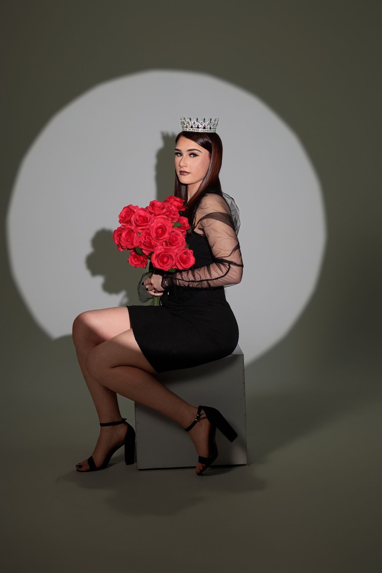 A model in a black dress poses on a cube with red roses and a crown in a dramatic spotlight setting.