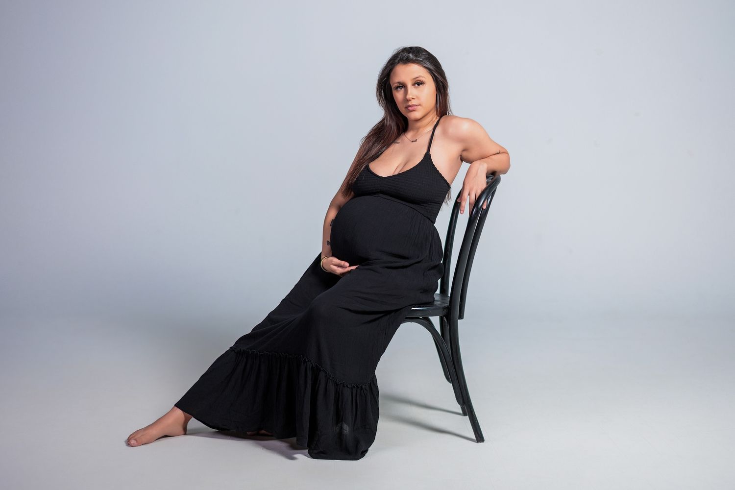 A person in a flowing black dress poses elegantly on a chair against a gray studio backdrop.