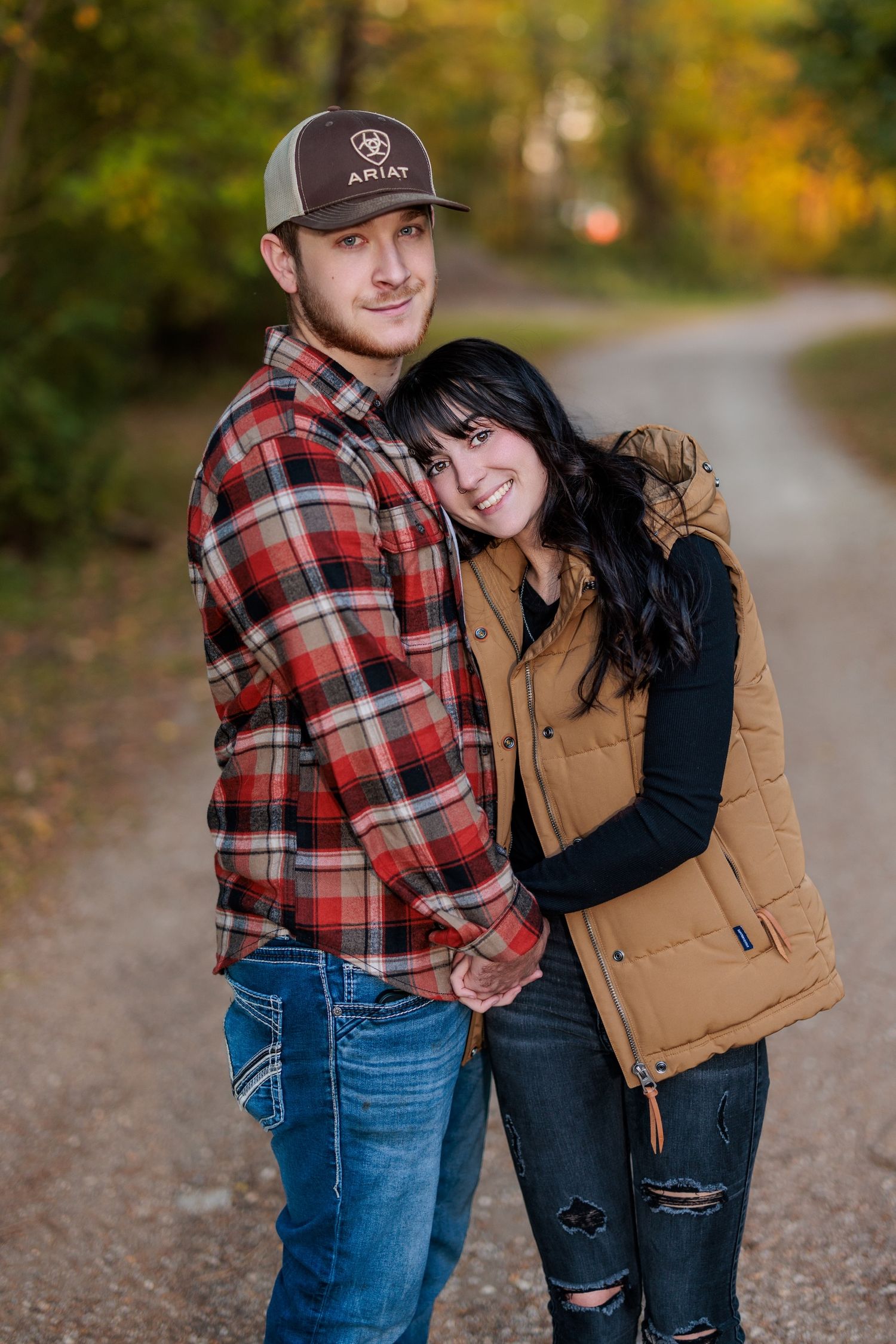 A couple in fall attire embraces on a dirt path surrounded by autumn foliage.