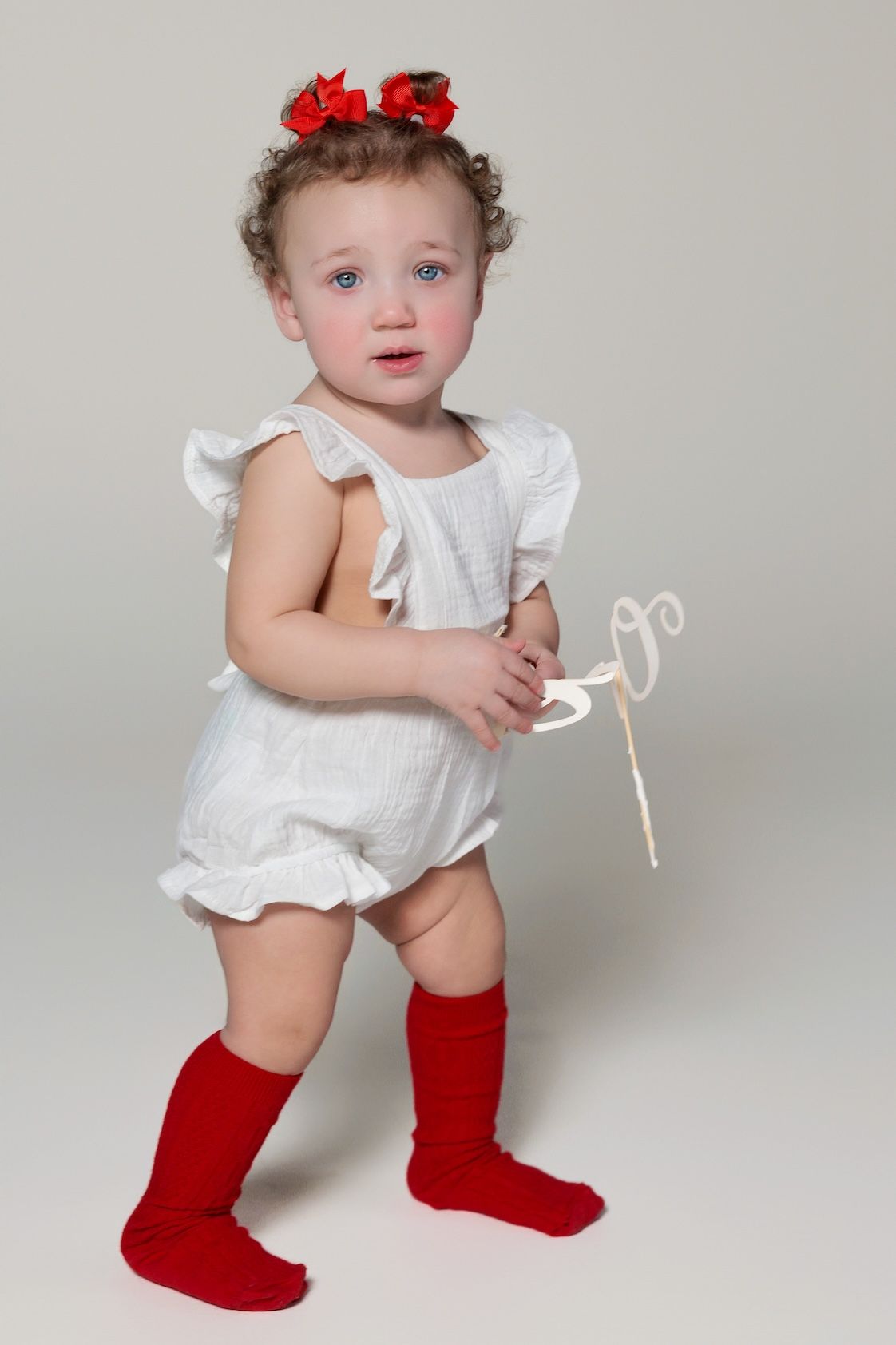 A toddler in a white ruffled romper and red knee socks stands against a gray backdrop holding a decorative wand.