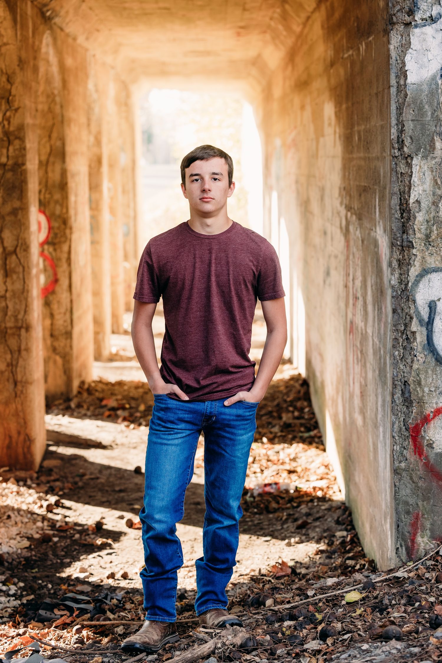 Figure stands in burgundy t-shirt and blue jeans inside an abandoned concrete tunnel with dramatic lighting.
