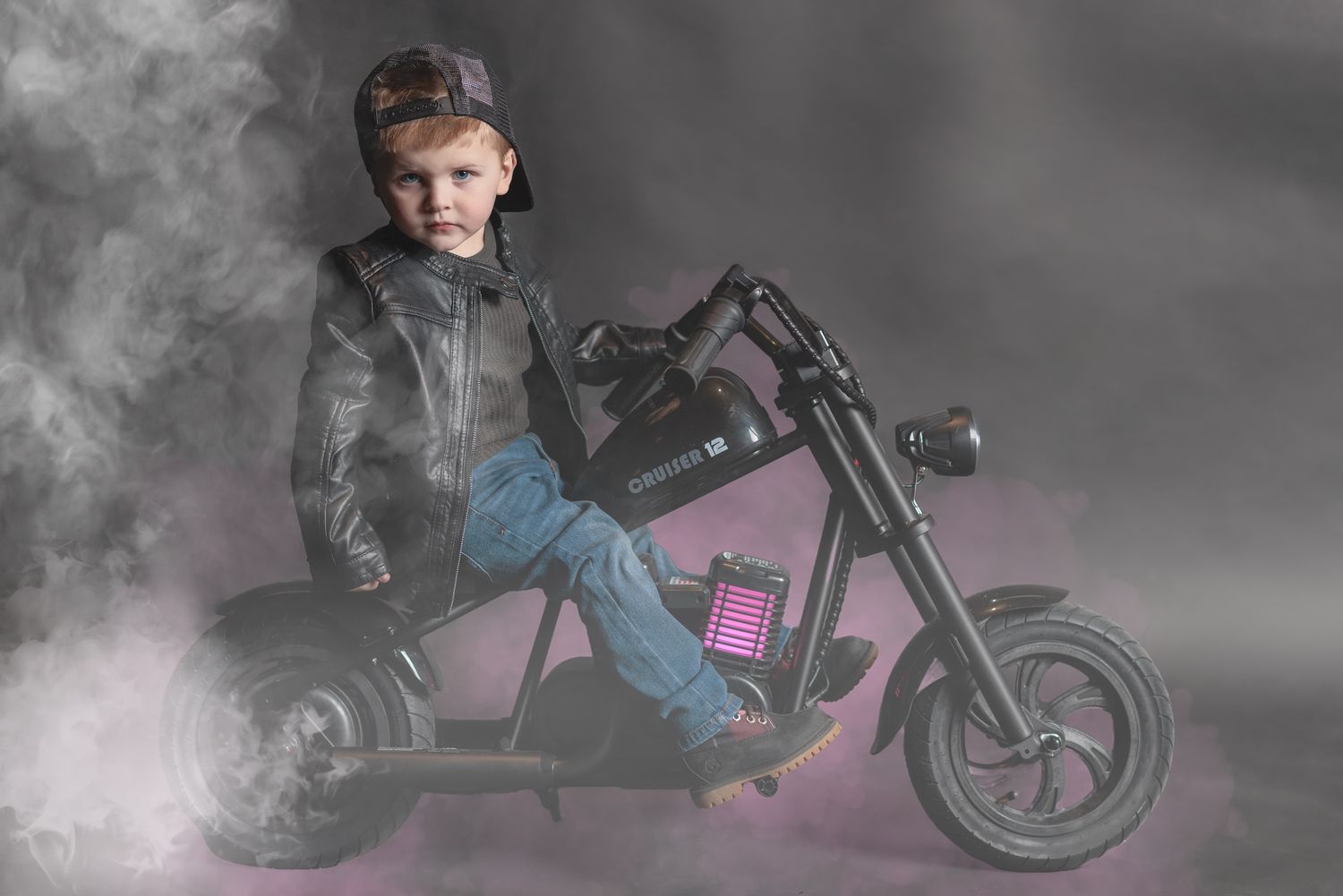 A small rider in leather jacket sits on a toy motorcycle amid dramatic fog and lighting effects.
