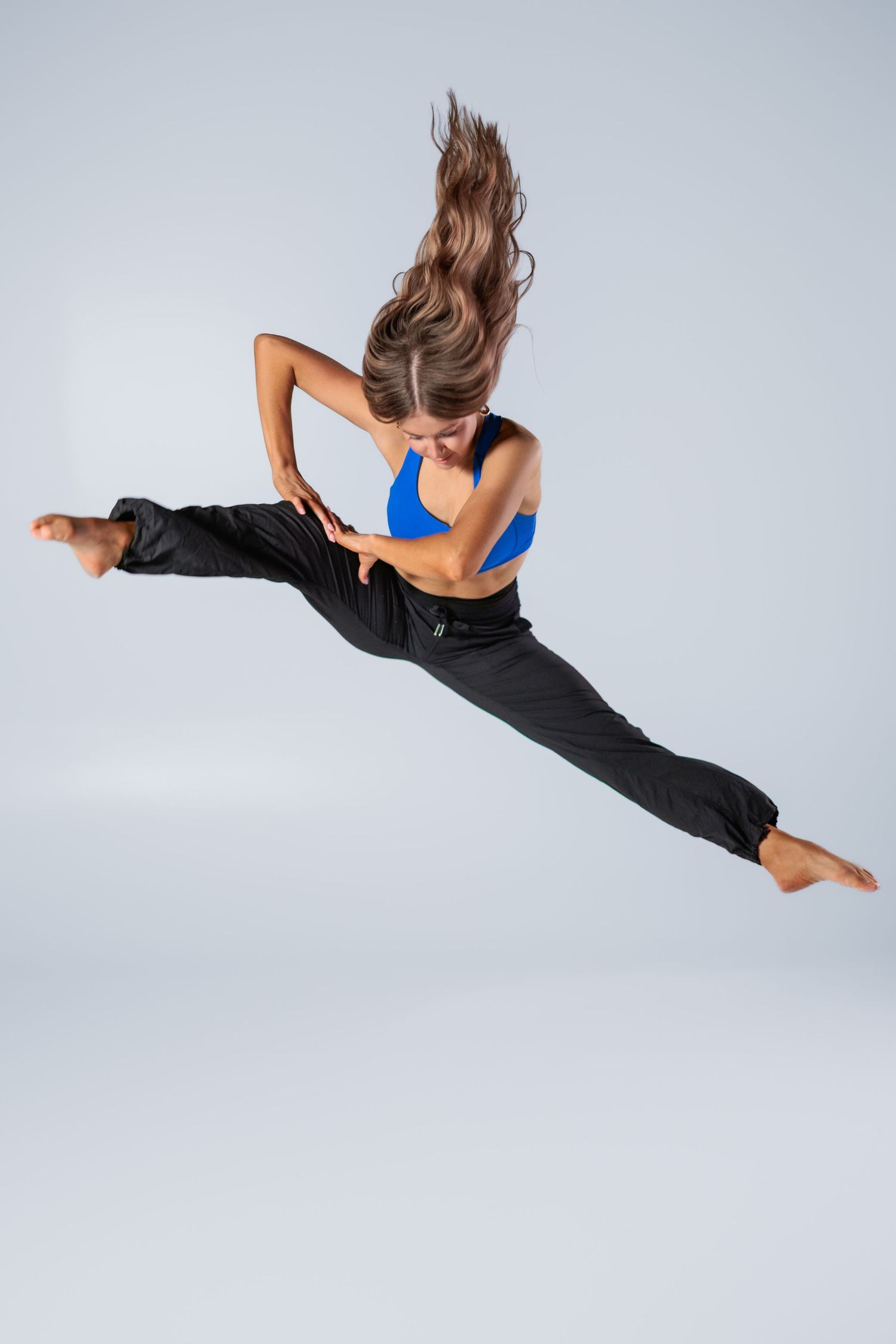 Athletic dancer in blue top and black pants performs dramatic split jump with flowing hair against gray background.