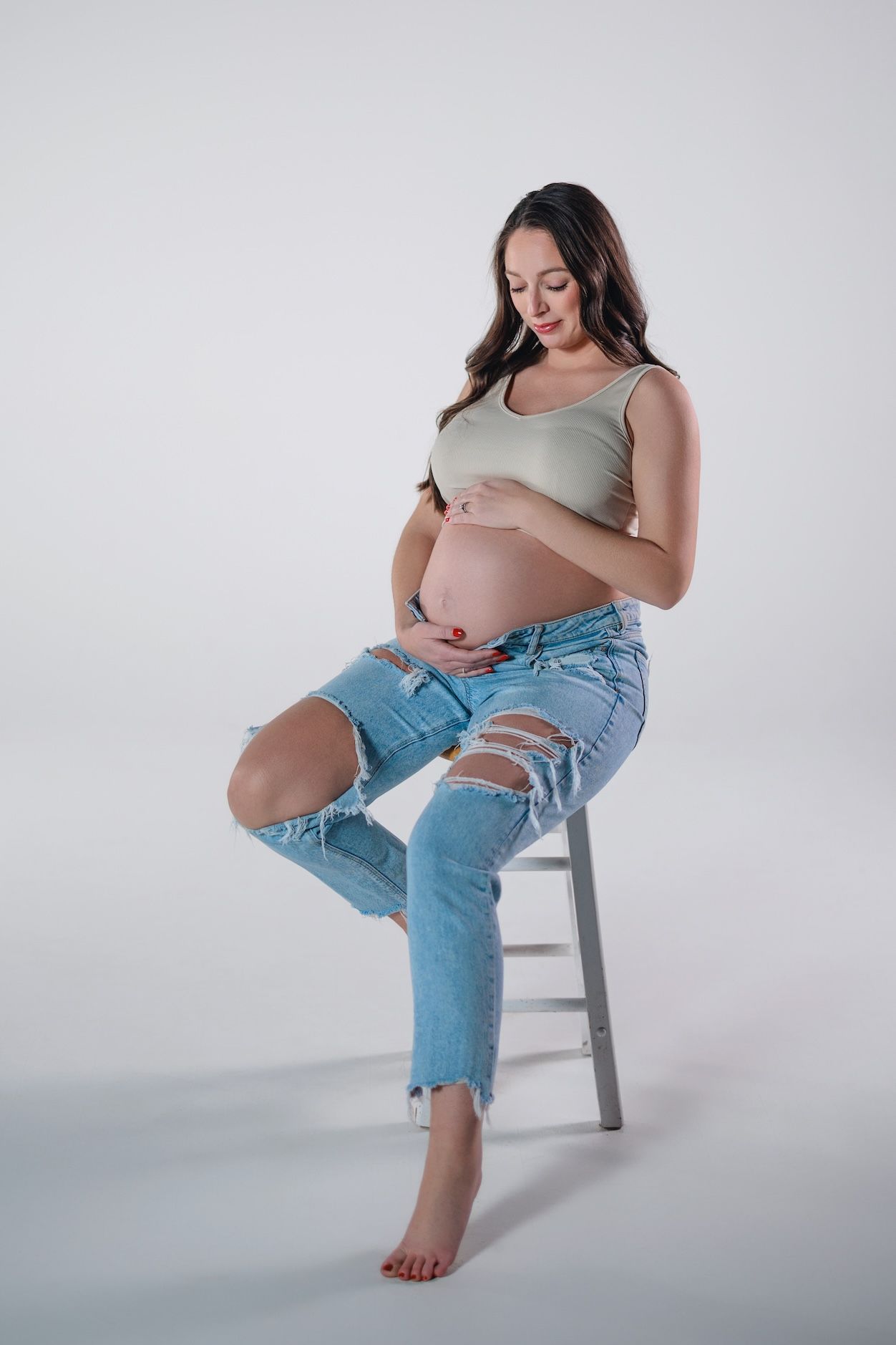 Expectant mother in gray tank top and distressed jeans sits casually on stool in studio setting.