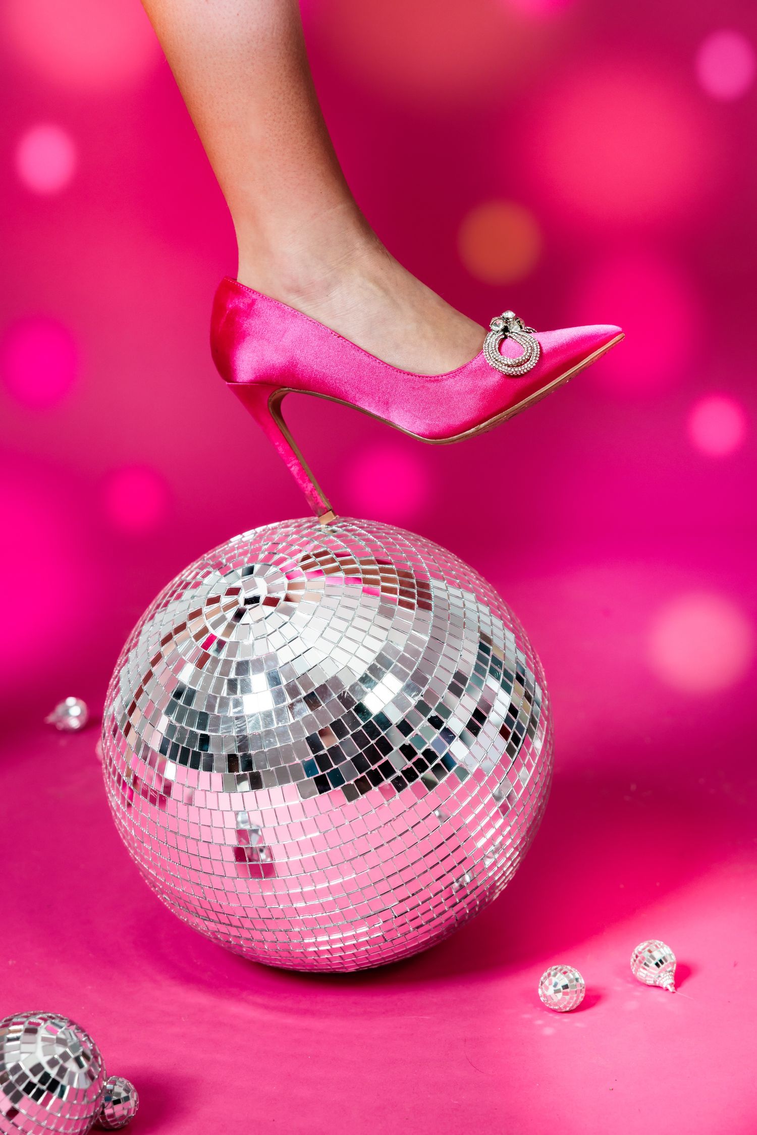 A pink stiletto heel rests elegantly on a silver disco ball against a vibrant pink background with sparkling bokeh effect.