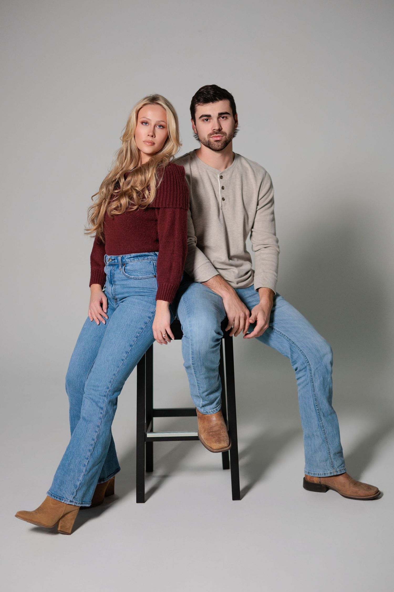 A couple wearing casual outfits with denim jeans pose together against a gray studio backdrop.