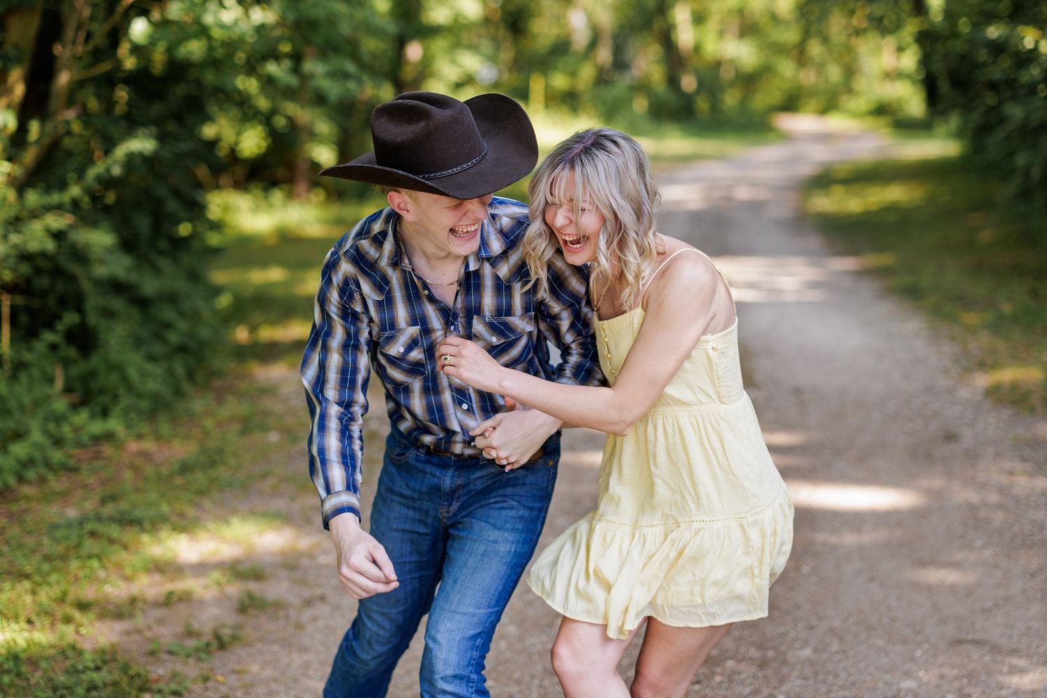 A couple walks down a sunlit path together, with one wearing a cowboy hat and plaid shirt while the other wears a yellow dress.