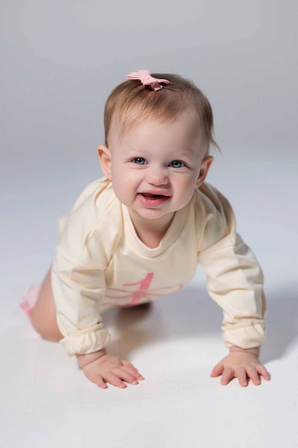 A happy baby in cream-colored clothing crawls on a white backdrop while wearing a small pink hair clip.