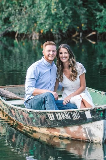 A couple sits together in a weathered canoe on calm waters surrounded by green foliage on a sunny day.