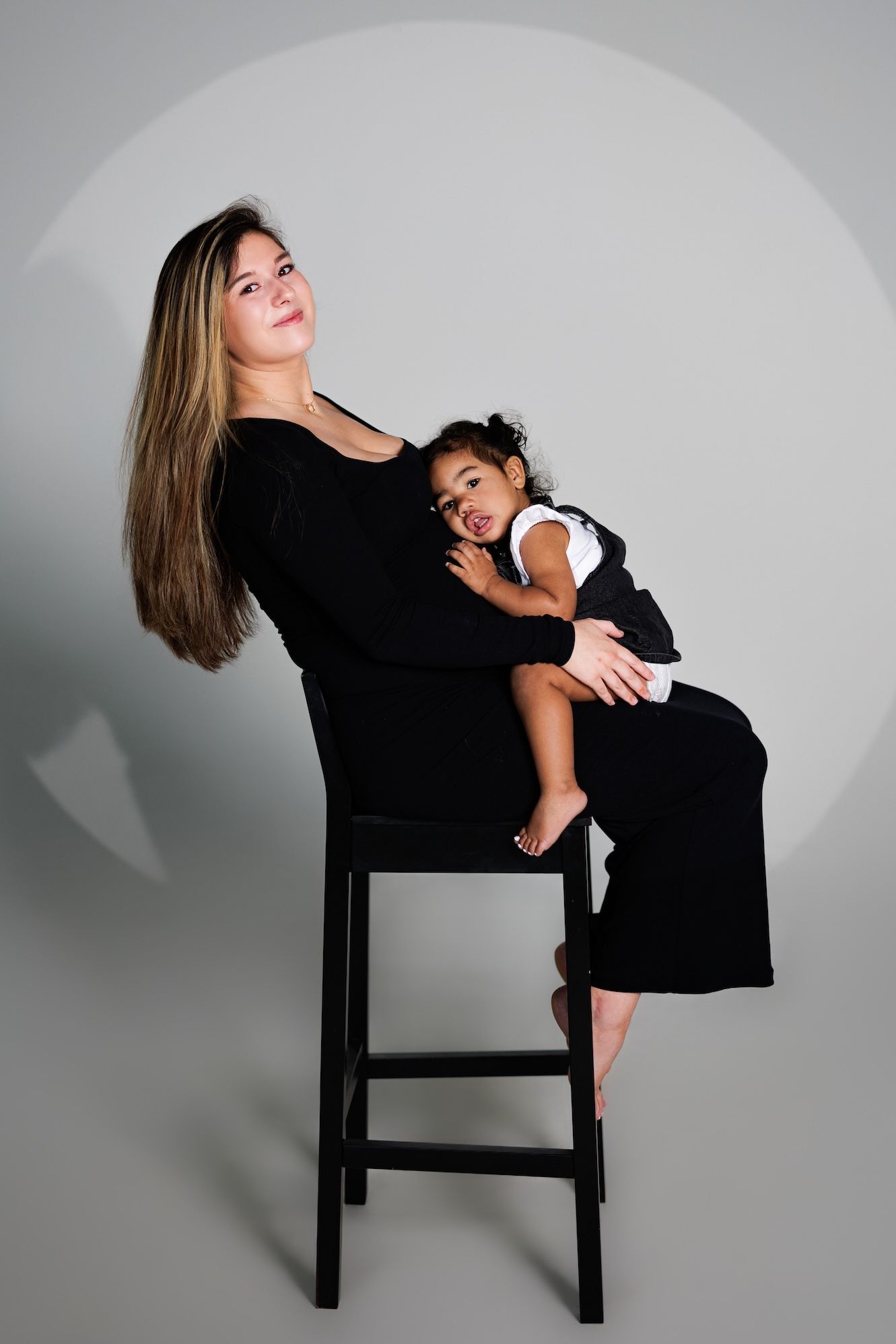 A mother and child share a tender moment on a black stool against a gray backdrop in a studio portrait setting.
