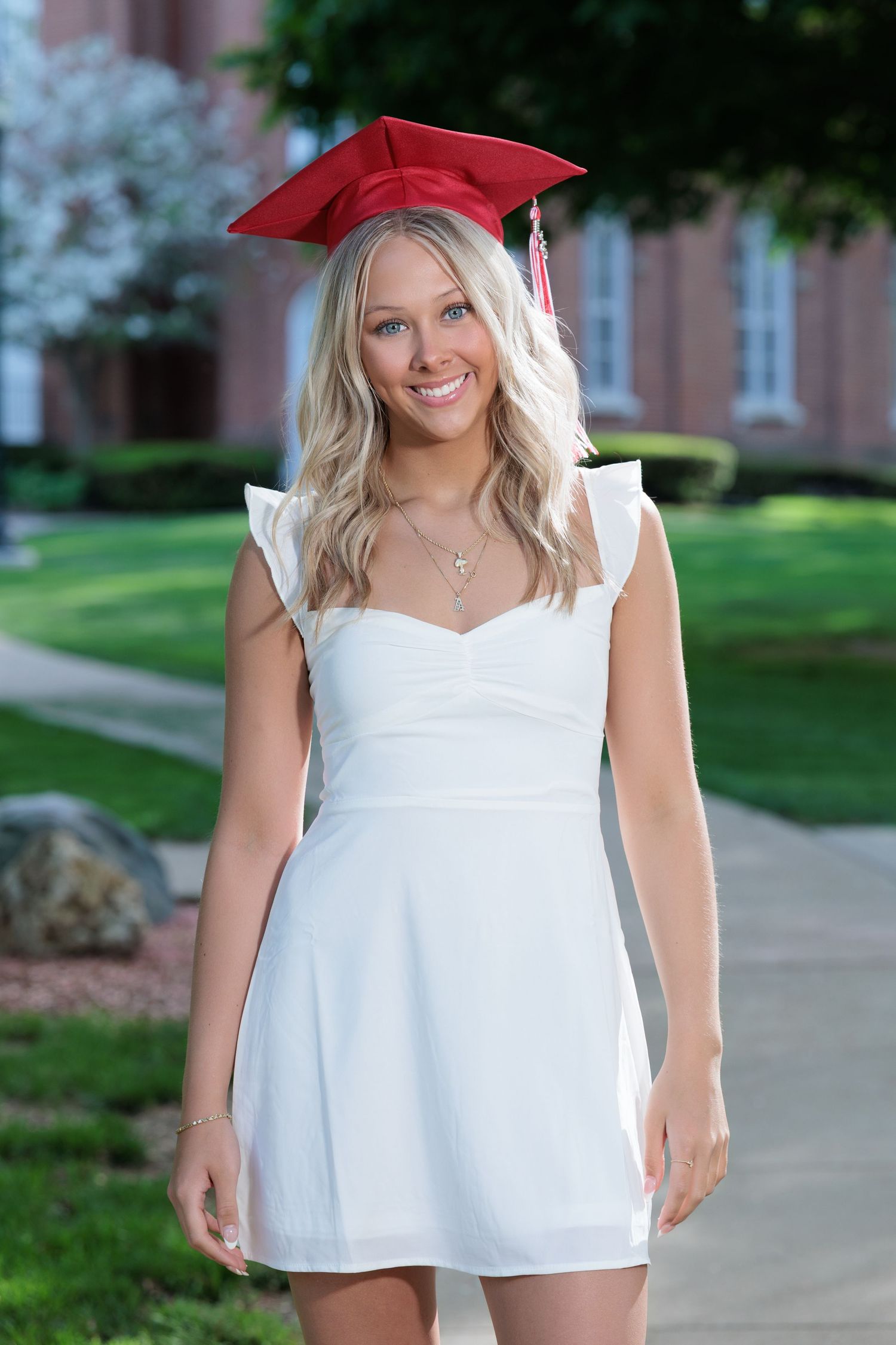 Graduate in red cap and white dress smiles while standing on campus sidewalk surrounded by greenery.