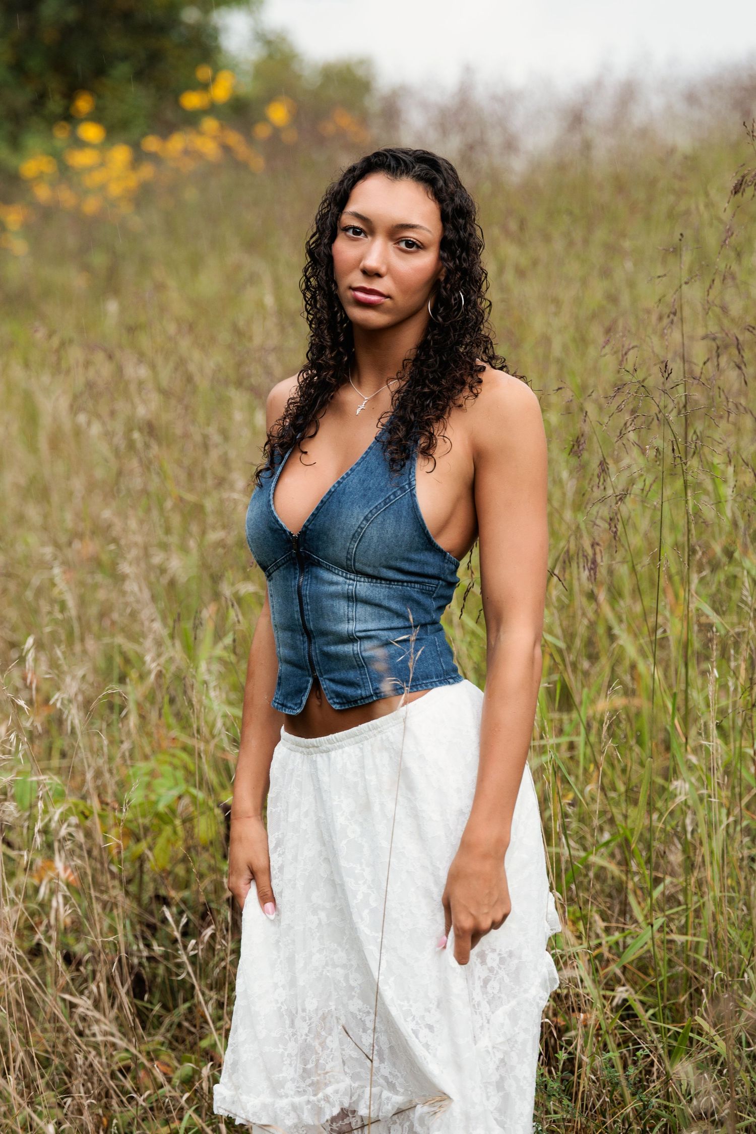 A person in a denim halter top and white lace skirt poses in a field of tall grass and wildflowers.