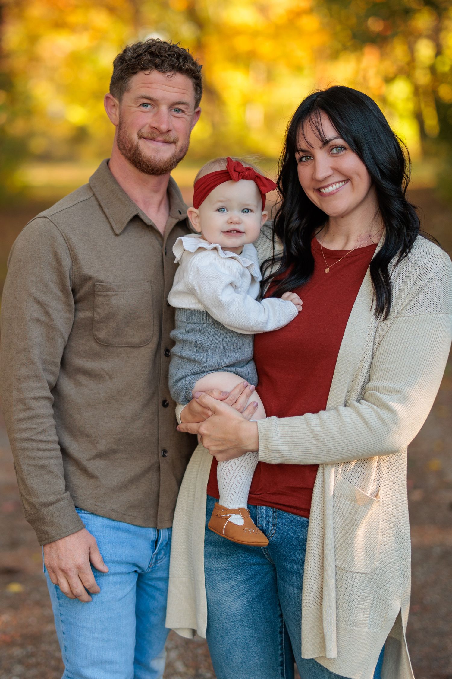 A family of three poses together outdoors on an autumn day, with golden fall foliage in the background.