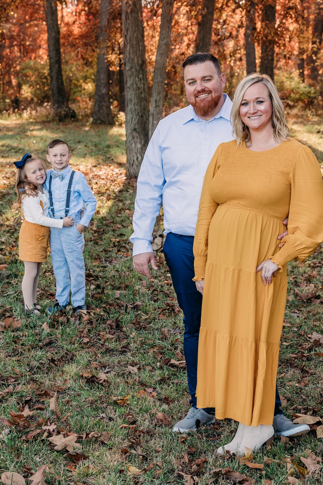 A family poses together in a wooded autumn setting wearing coordinated mustard yellow and light blue outfits.