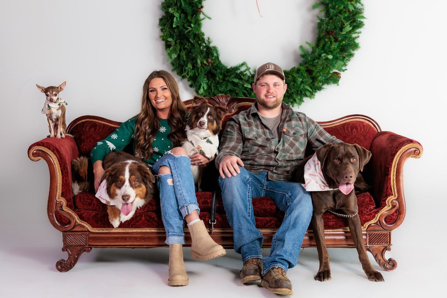 Family portrait on vintage red couch with three dogs and holiday wreath decor in background.