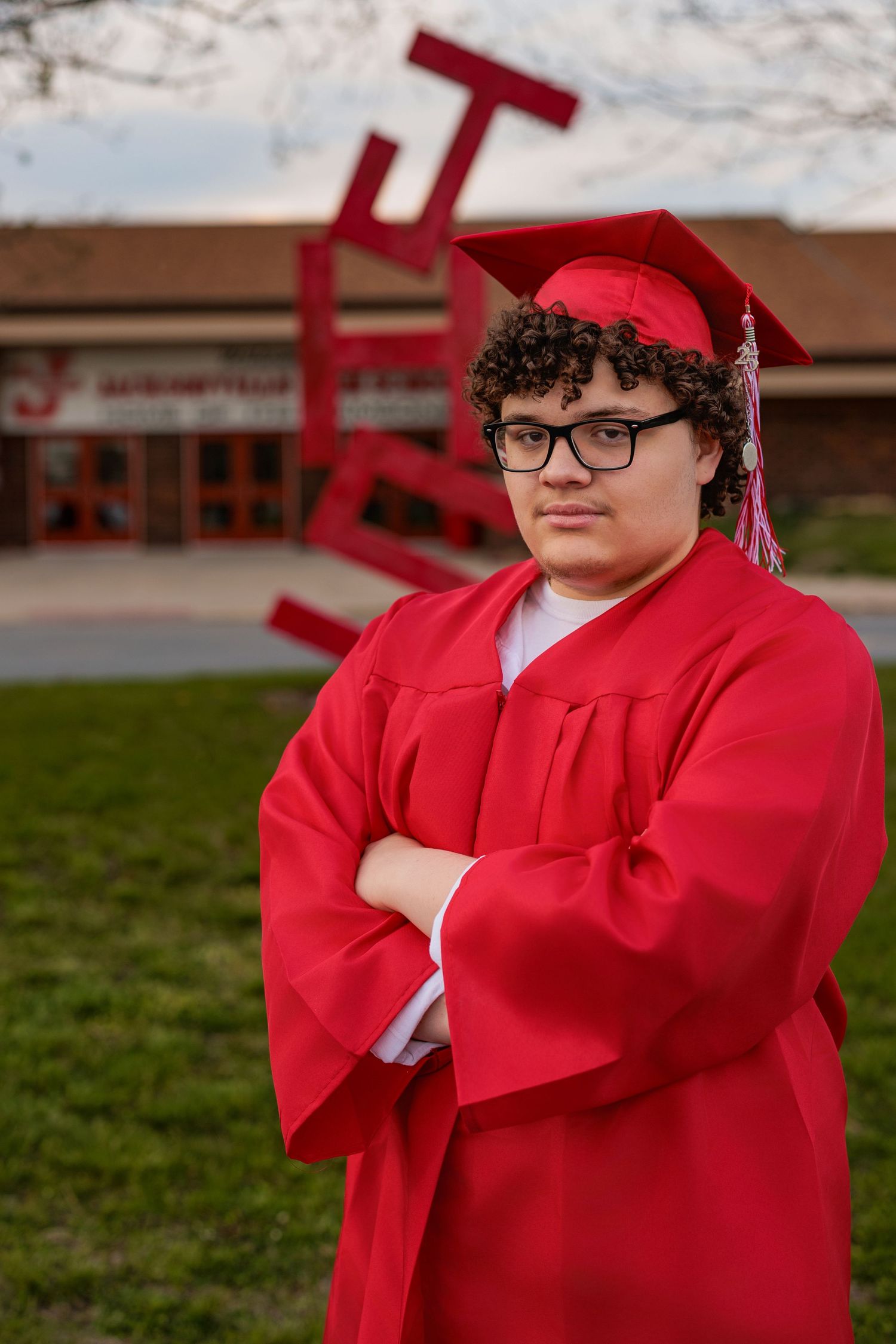 Graduation photo of student in red cap and gown standing with crossed arms in front of campus building and sculpture.