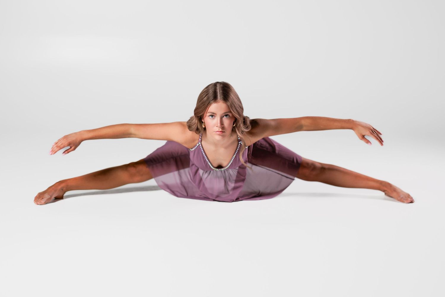 Dancer in purple leotard performs split stretch pose on white studio floor.