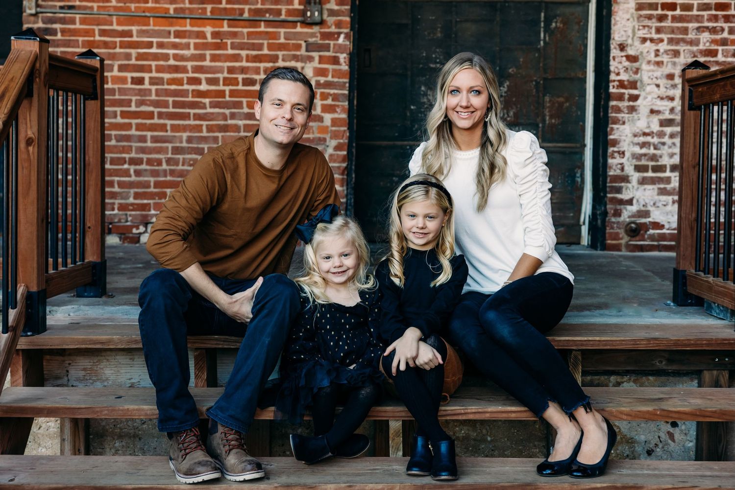 Family of four sits together on brick building steps wearing coordinated casual outfits for a portrait.