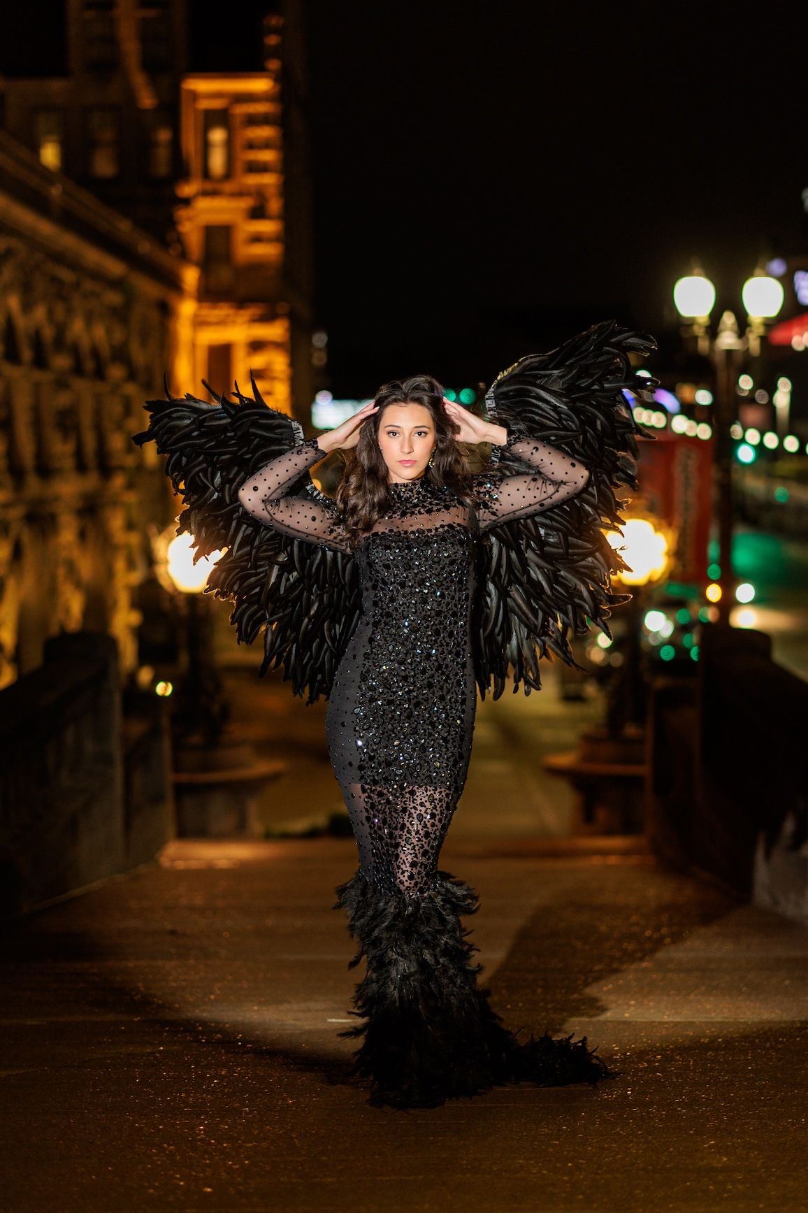 Dramatic black feathered dress with lace detail photographed at night against illuminated building and street lights.