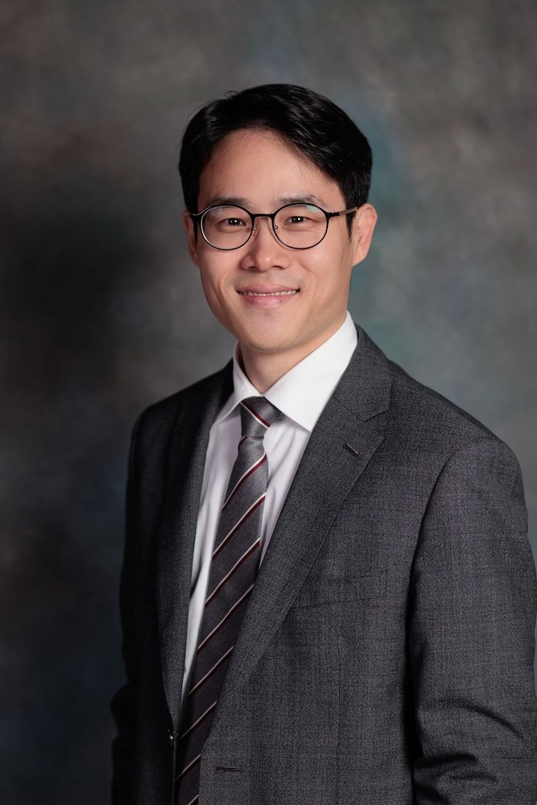 Professional headshot portrait of someone wearing glasses, a dark gray suit and striped tie against a mottled background.
