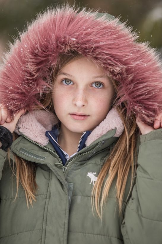 Winter portrait in an olive green puffer coat with pink fur-trimmed hood and striking blue eyes.