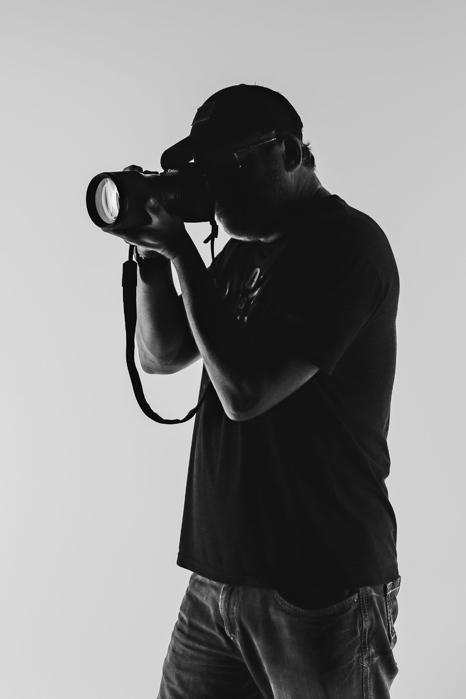 Silhouette of photographer in black t-shirt taking photo with DSLR camera against light gray background.