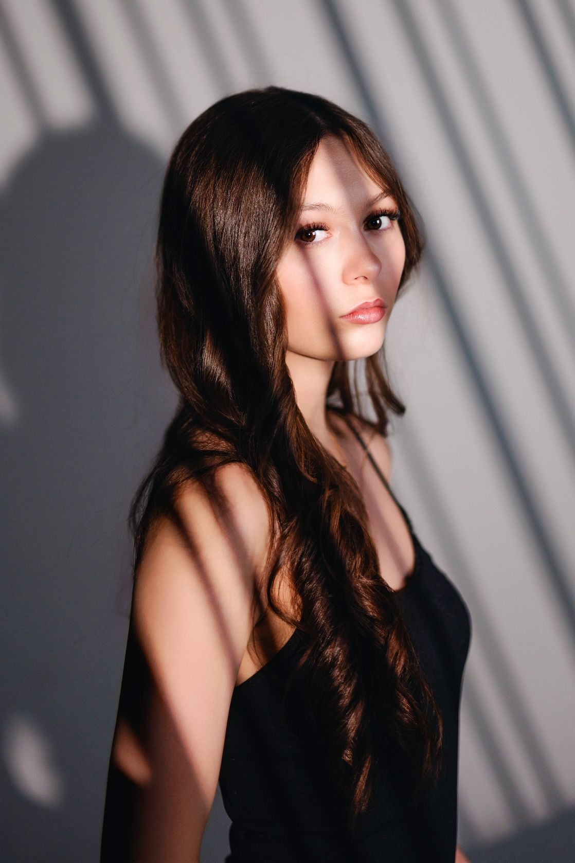 Artistic portrait with dramatic diagonal shadows on wall featuring long dark hair and black sleeveless top.