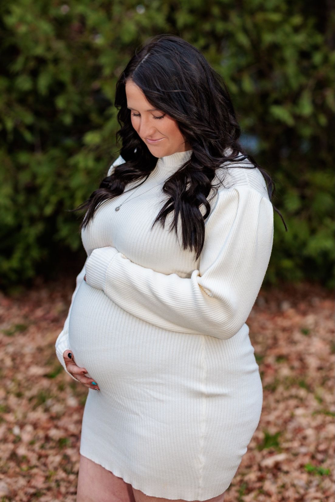A pregnant woman in a white sweater dress poses outdoors against a green backdrop of foliage.
