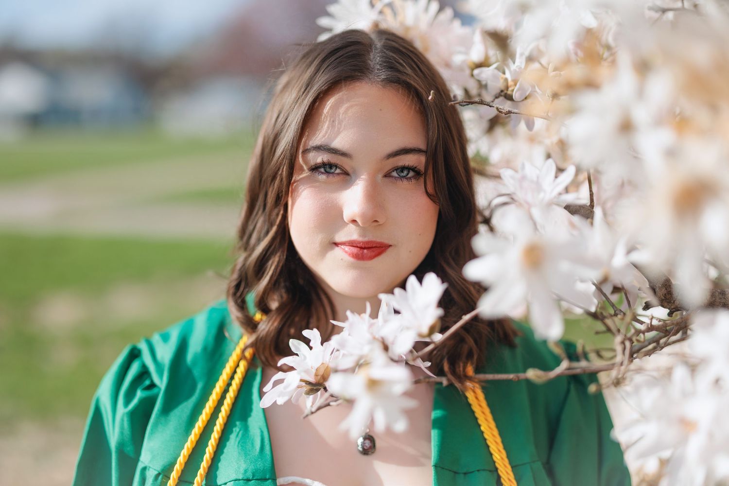 A graduate in emerald green cap and gown poses with white flowers on a spring day.