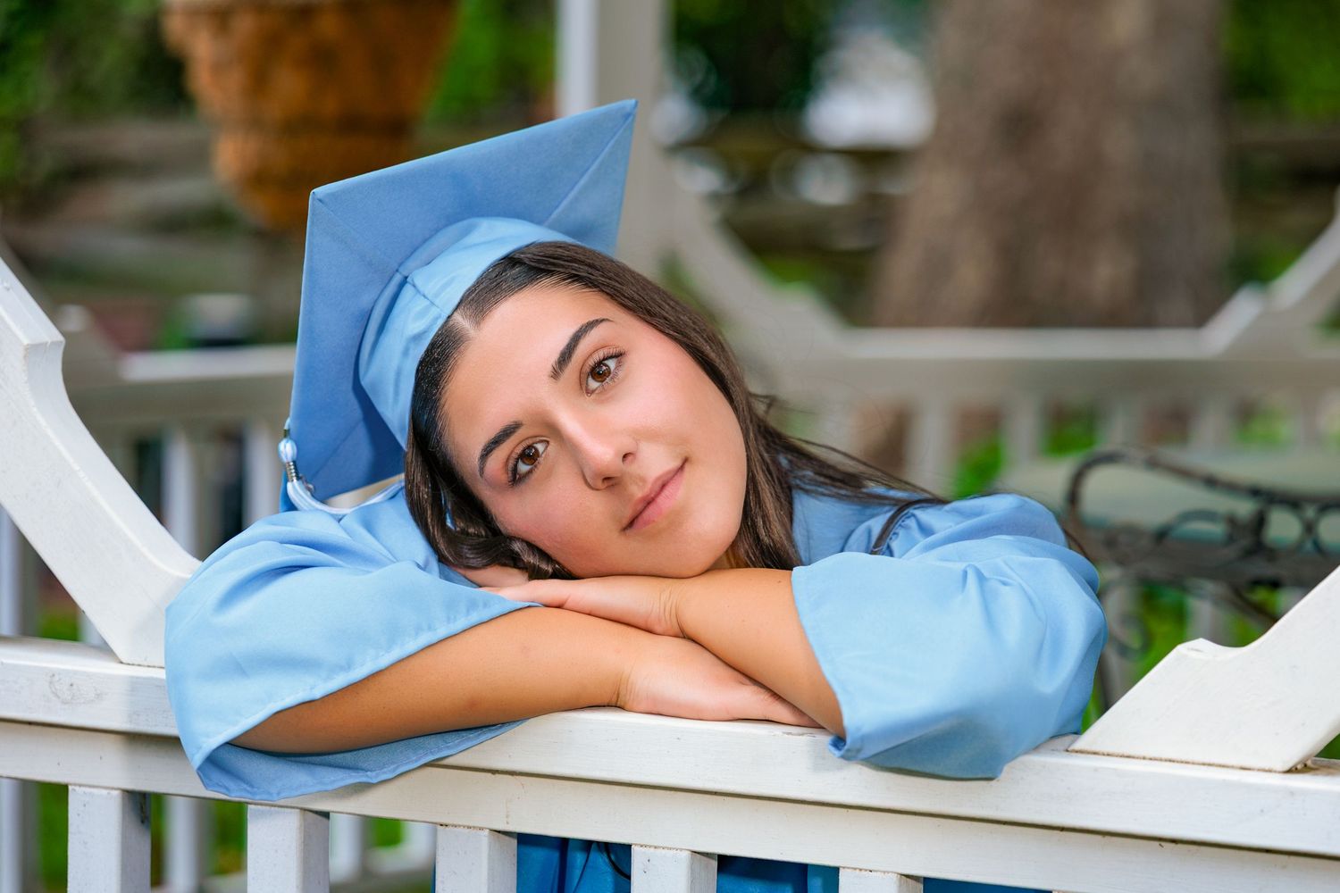 Graduate in light blue cap and gown rests head on hands while posing on white railing outdoors.