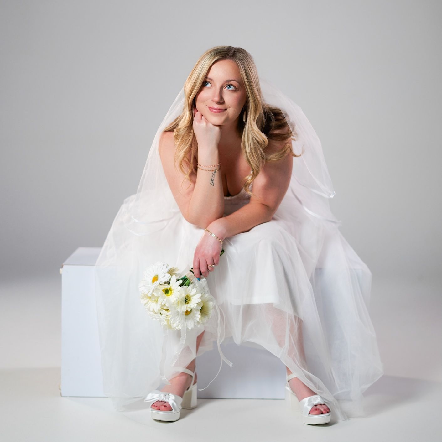 A bride in a white wedding dress and veil sits on a cube holding a bouquet of white flowers.