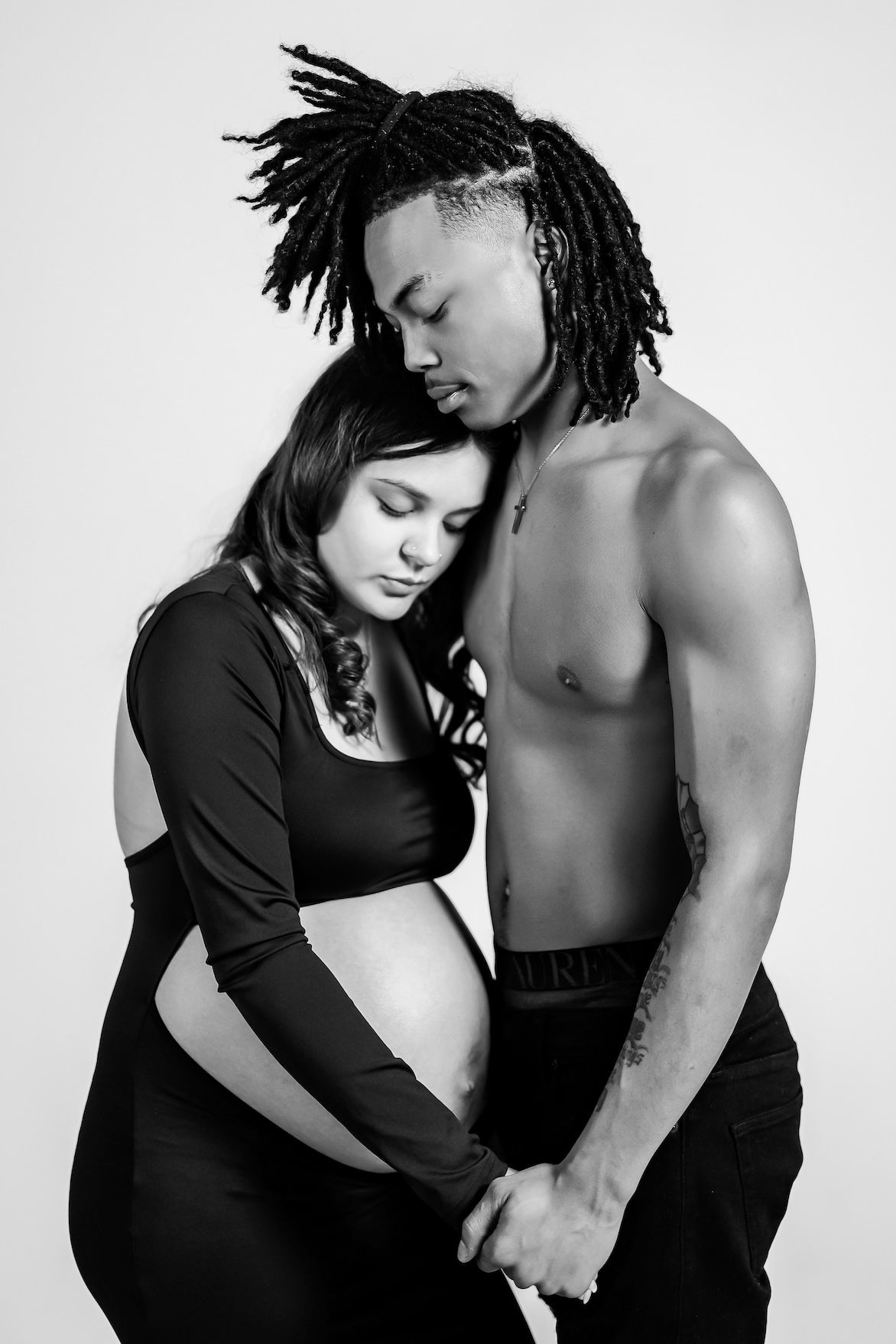 Black and white intimate maternity photo showing a couple embracing with woman in black top and shirtless muscular partner.
