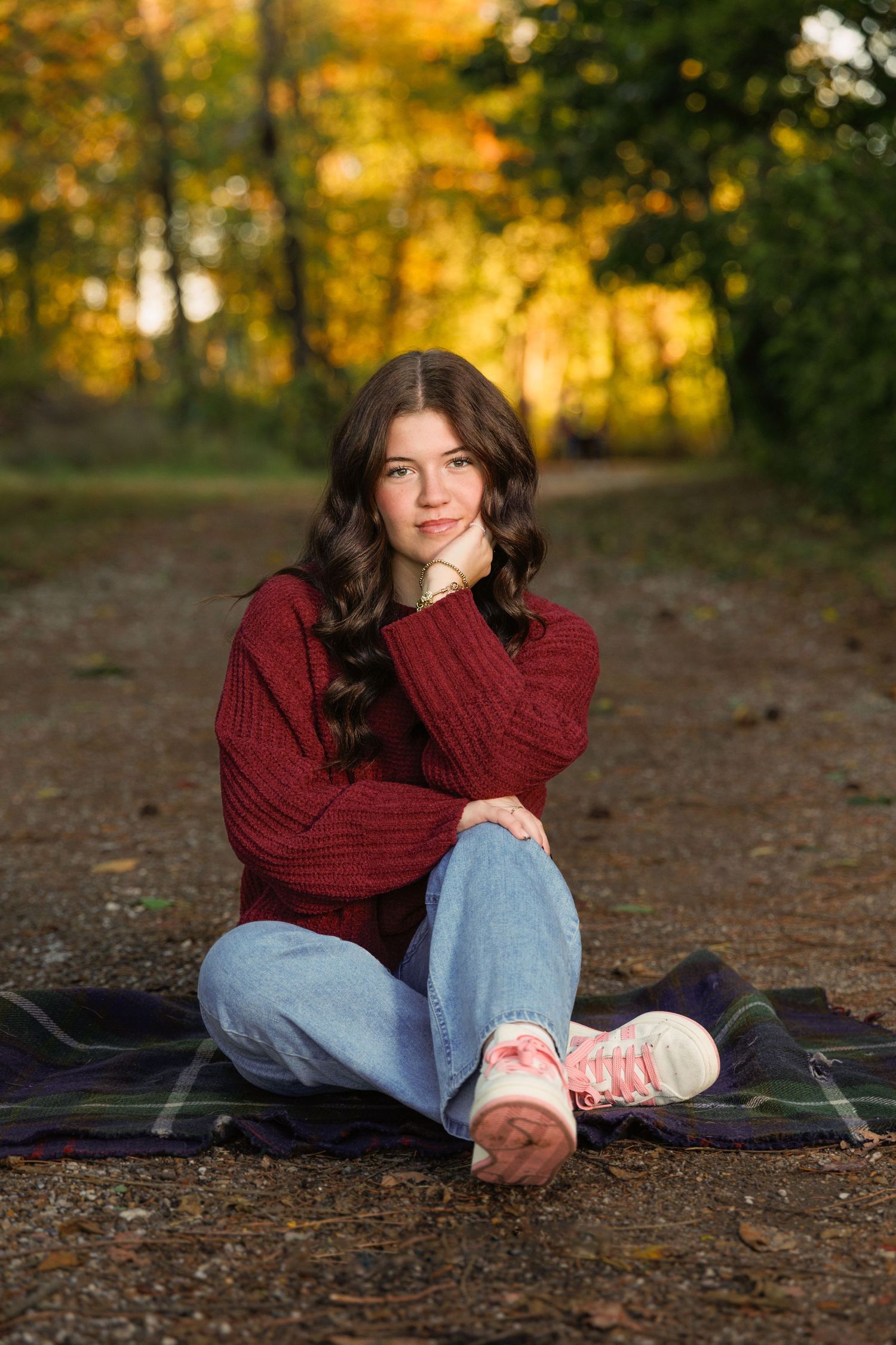 A person in a burgundy sweater and jeans sits on a blanket in the woods during autumn with golden foliage in background.
