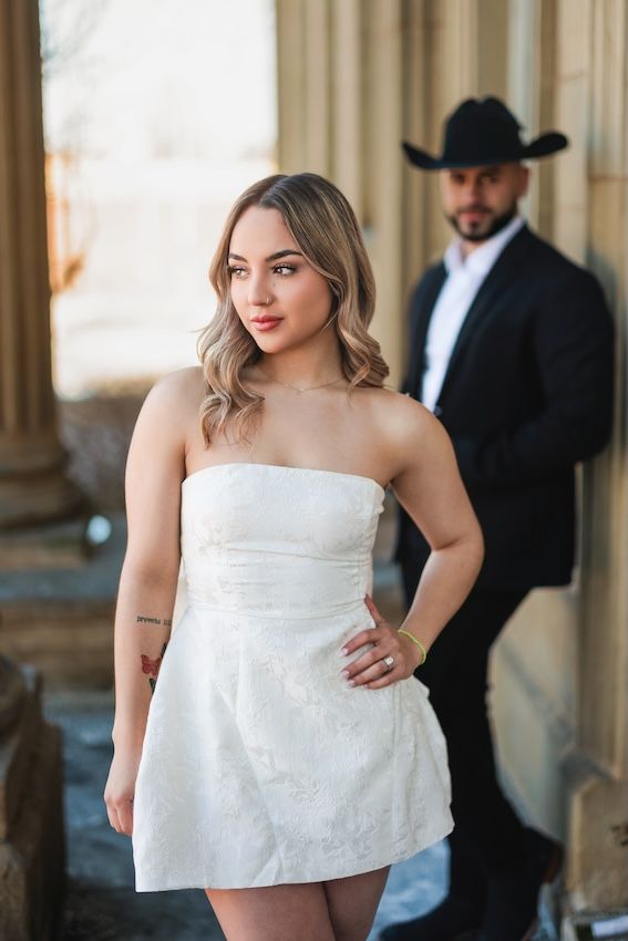 A couple in a rustic setting with a white strapless dress and cowboy attire pose near stone pillars.