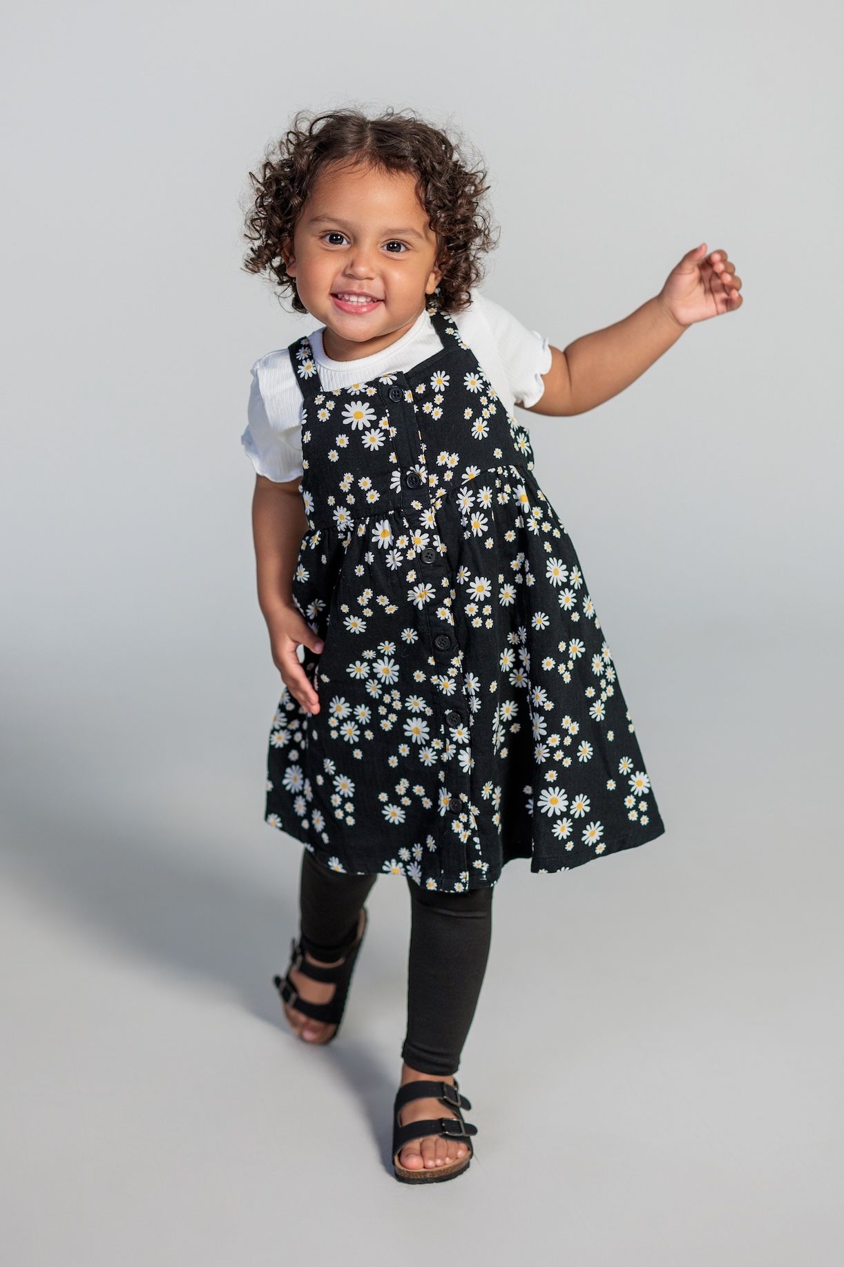 Child wearing a floral patterned black dress over leggings and sandals poses against a grey background.