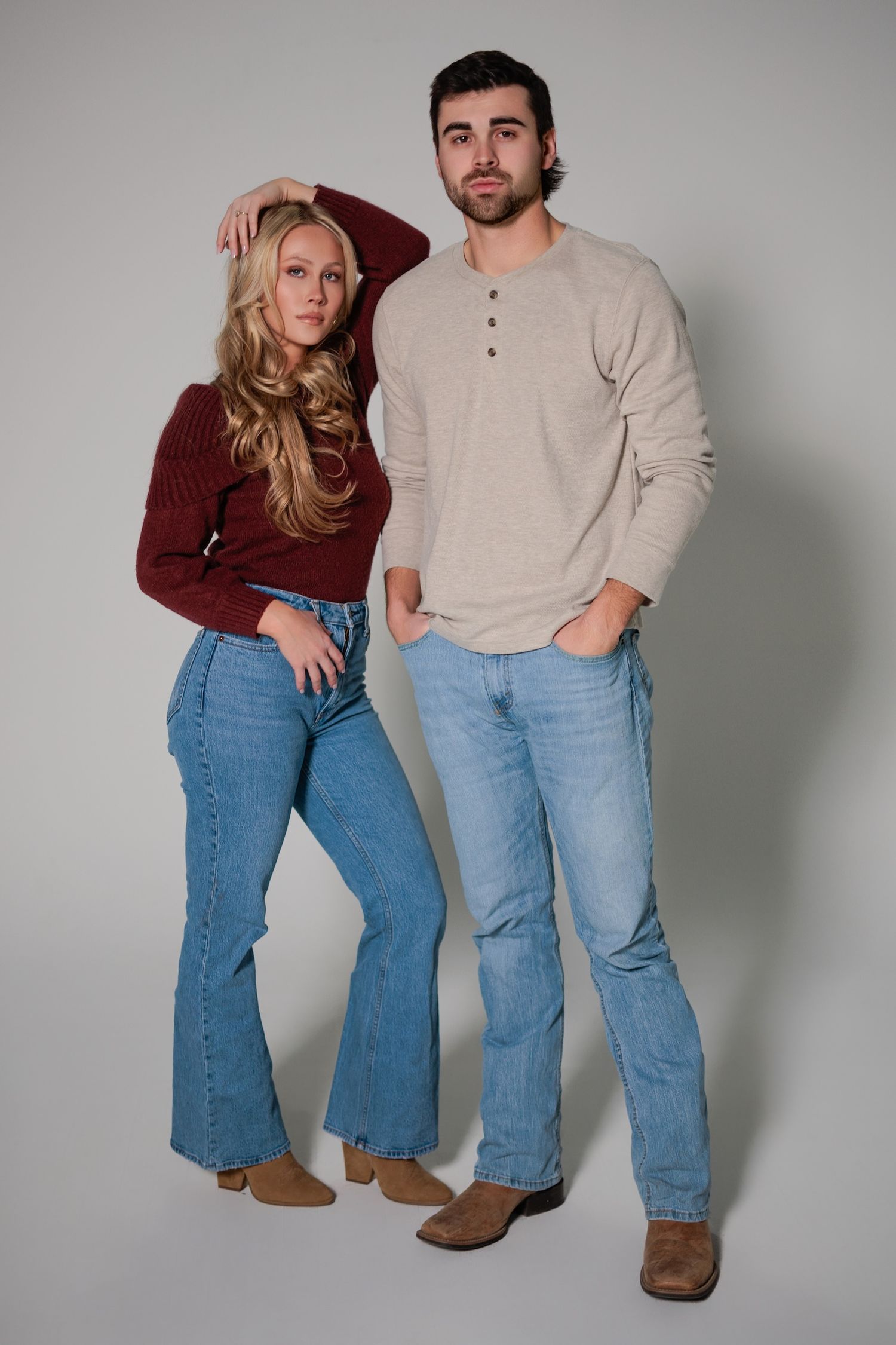 Two people pose in casual attire with burgundy sweater and beige henley paired with light blue jeans against grey backdrop.