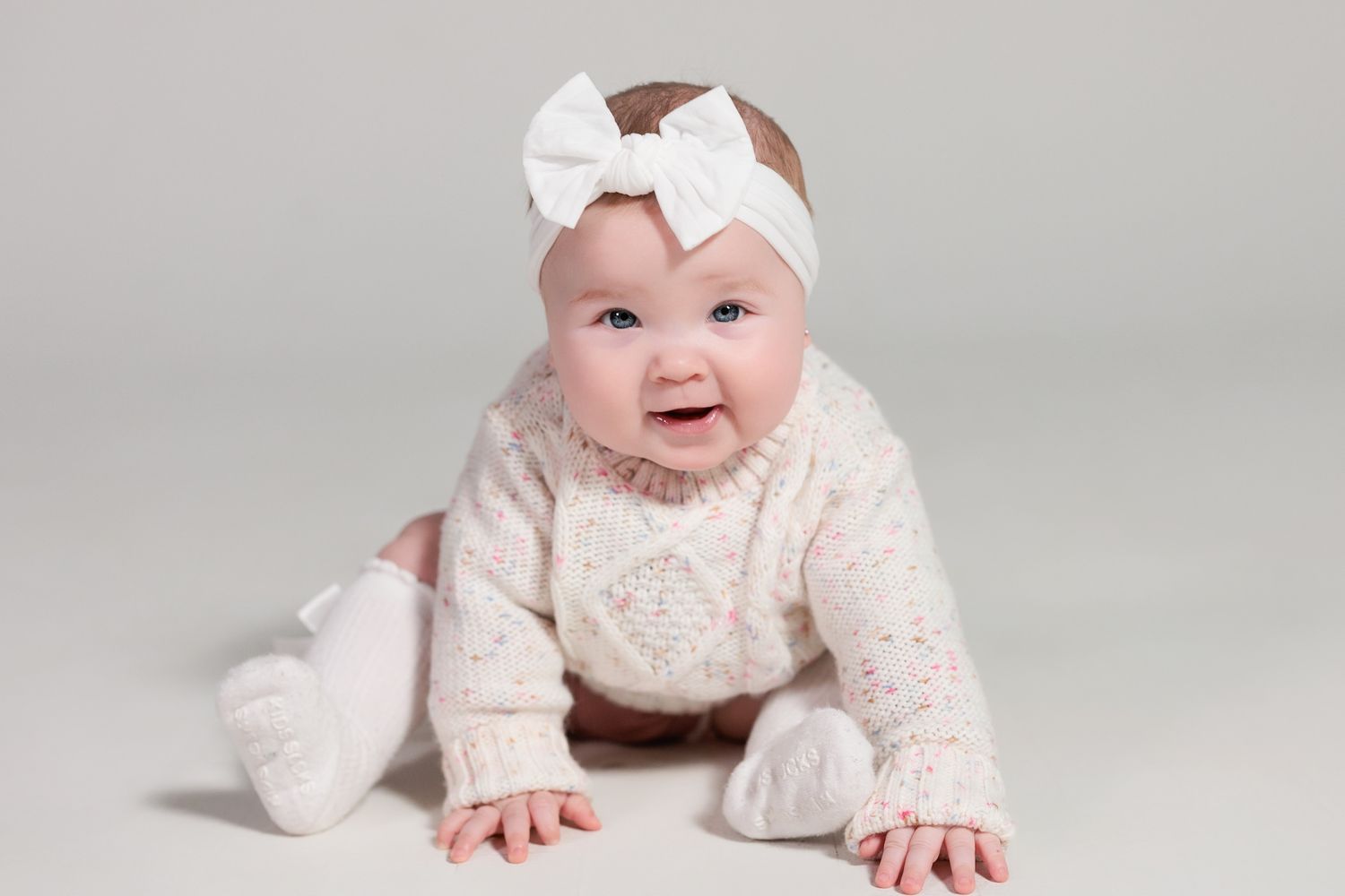 An infant wearing a white headbow and lace dress lies on their stomach on a light gray backdrop.