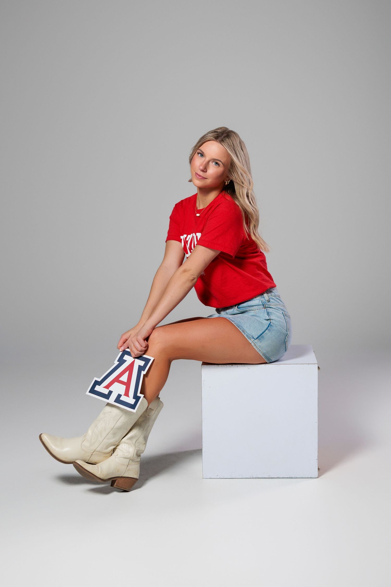 Model in red shirt and denim shorts poses on white cube with cowboy boots featuring collegiate A logo.